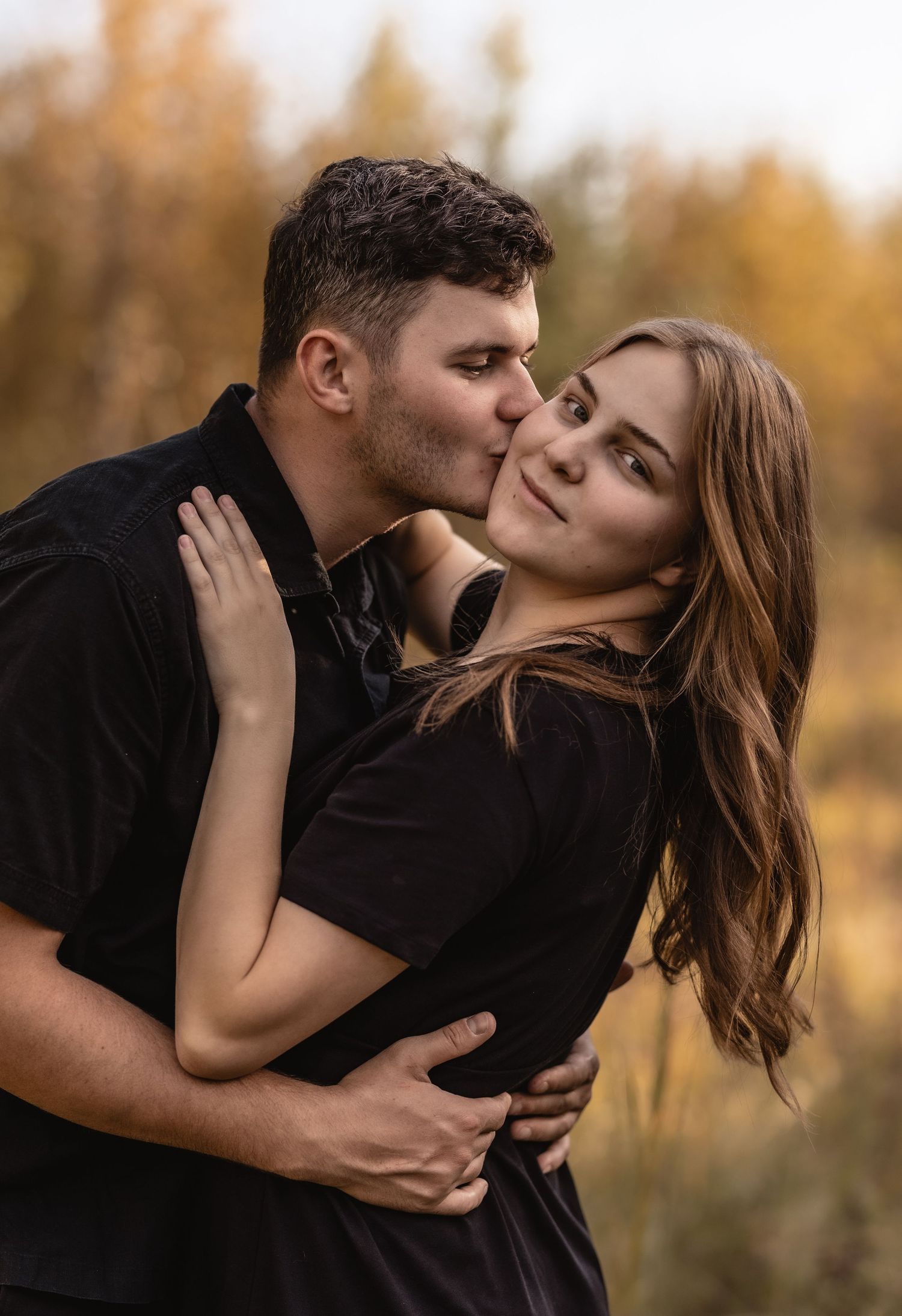 Couple in matching black outfits embracing outdoors, man kissing woman on cheek with autumn foliage background.