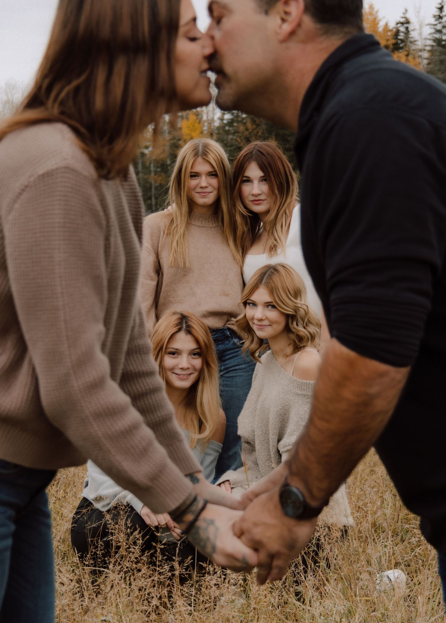 Family portrait of parents kissing while three daughters watch in a golden autumn meadow.