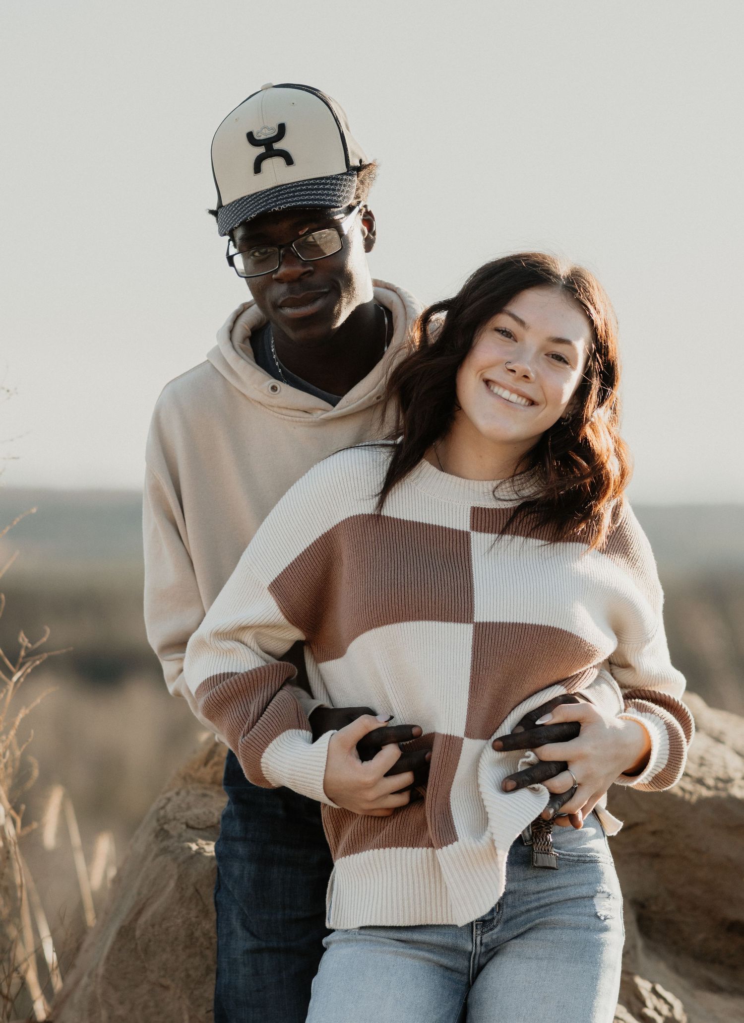 Couple embracing outdoors in casual sweaters, smiling at camera during golden hour in a scenic landscape.