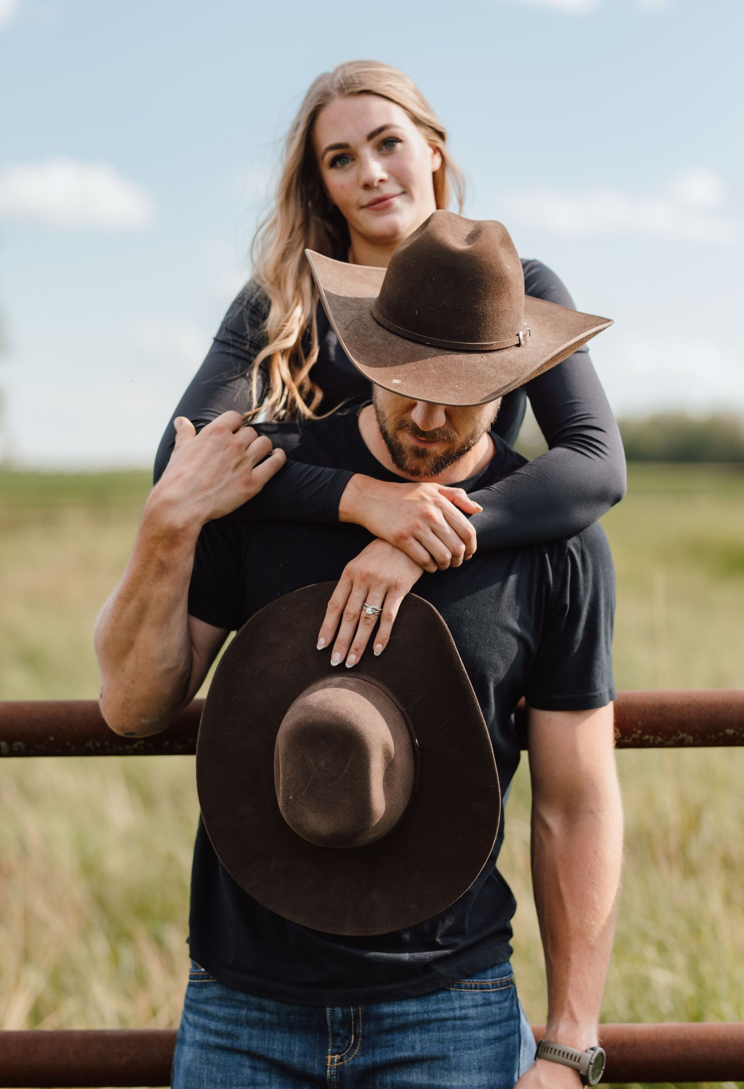 Couple posing outdoors near a rustic fence, woman embracing man wearing a cowboy hat in a green field.