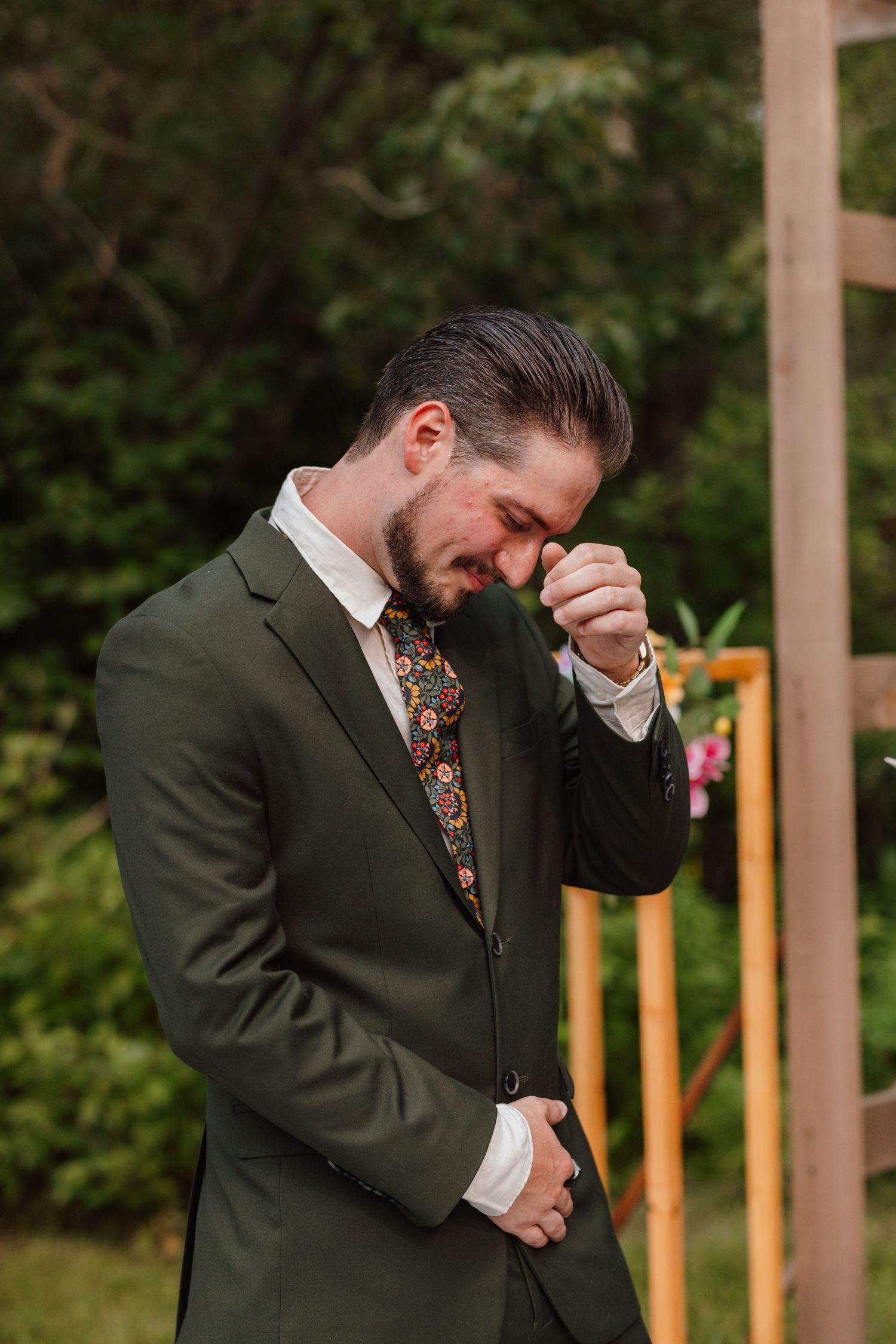 Emotional groom in olive green suit with floral tie wiping tears at outdoor wedding ceremony.