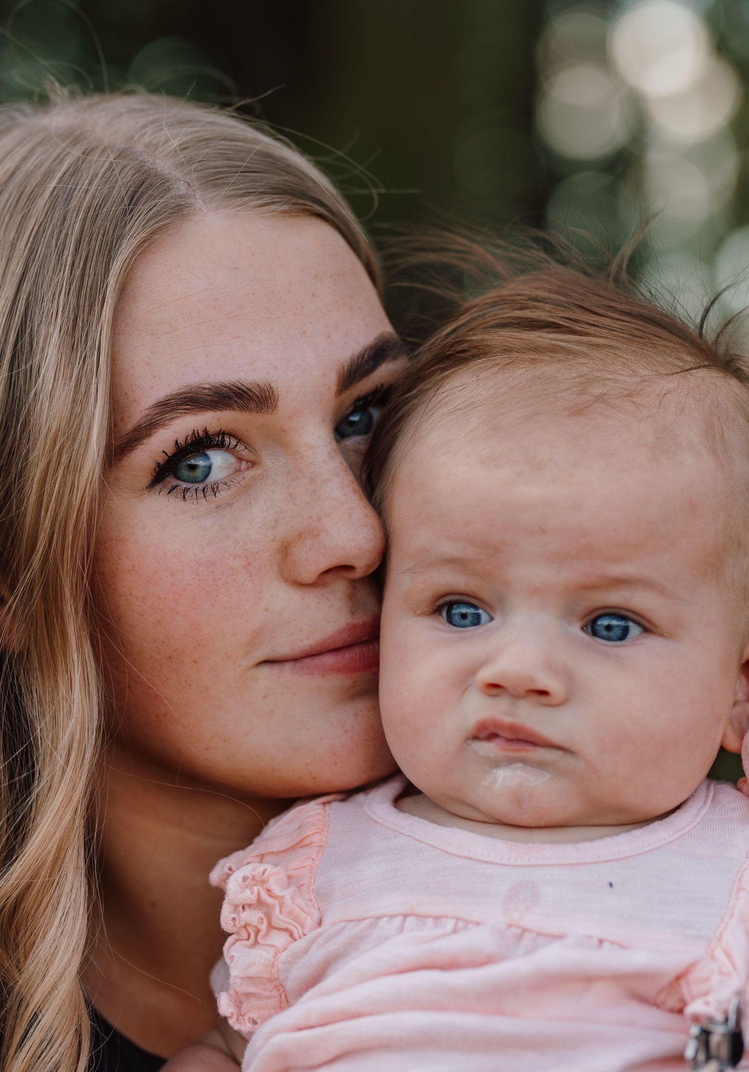 A young blonde woman with blue eyes holds a blue-eyed baby in a pink outfit, both facing the camera outdoors.