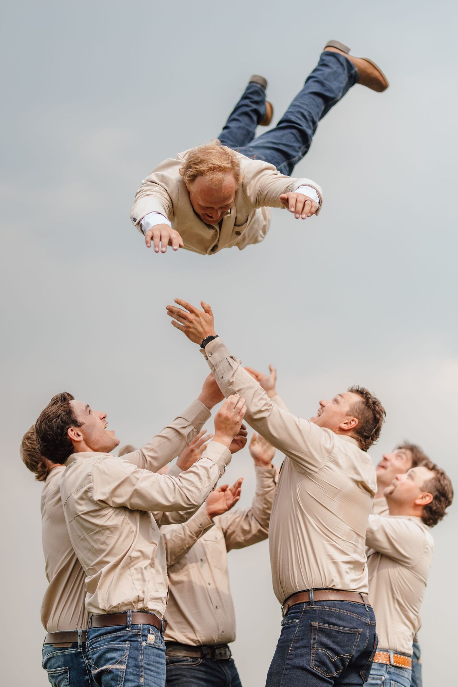 Groomsmen tossing the groom into the air in celebration against a cloudy sky backdrop.