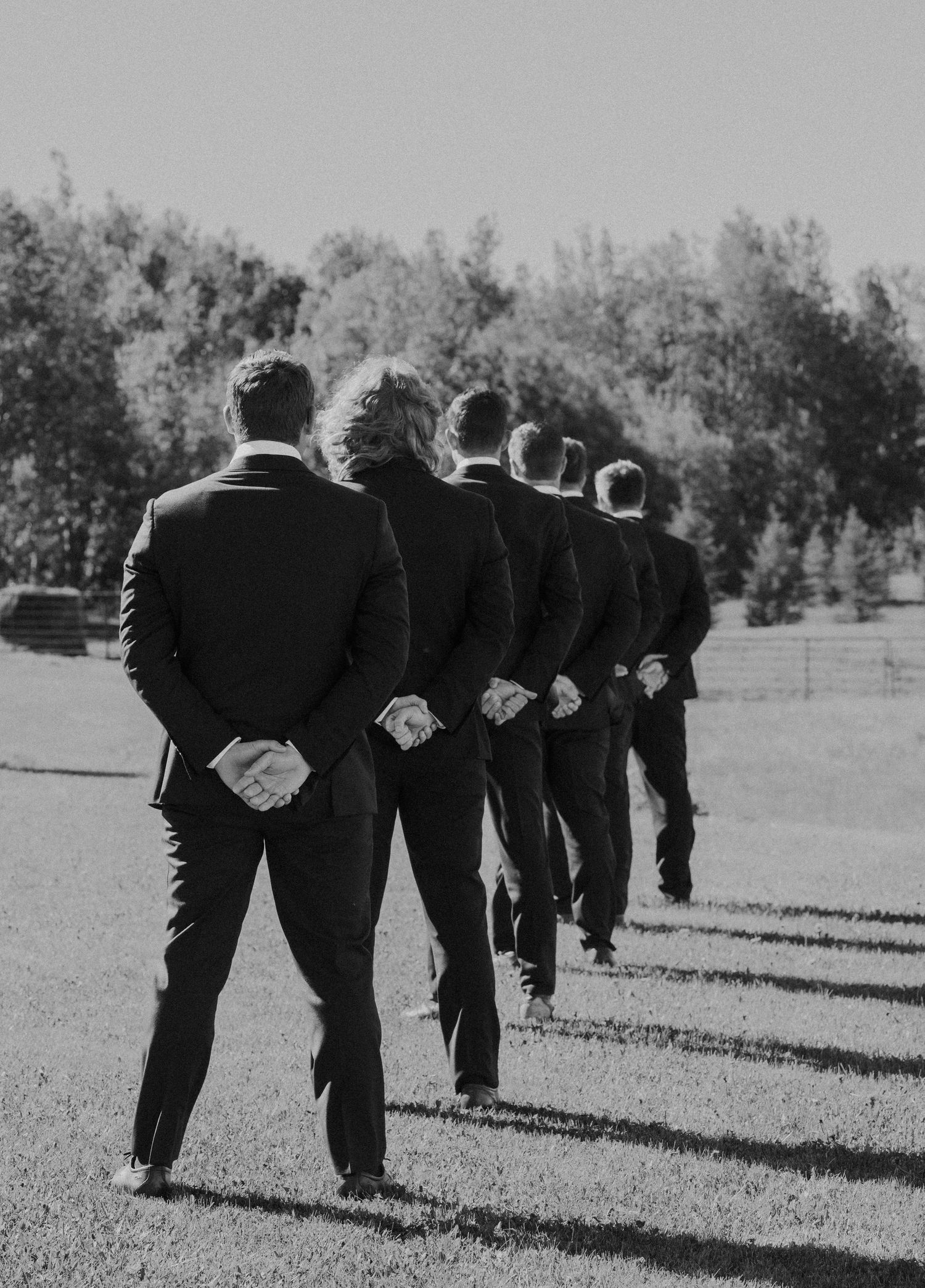 Black and white photo of groomsmen in suits walking together outdoors on a sunny day at a wedding.