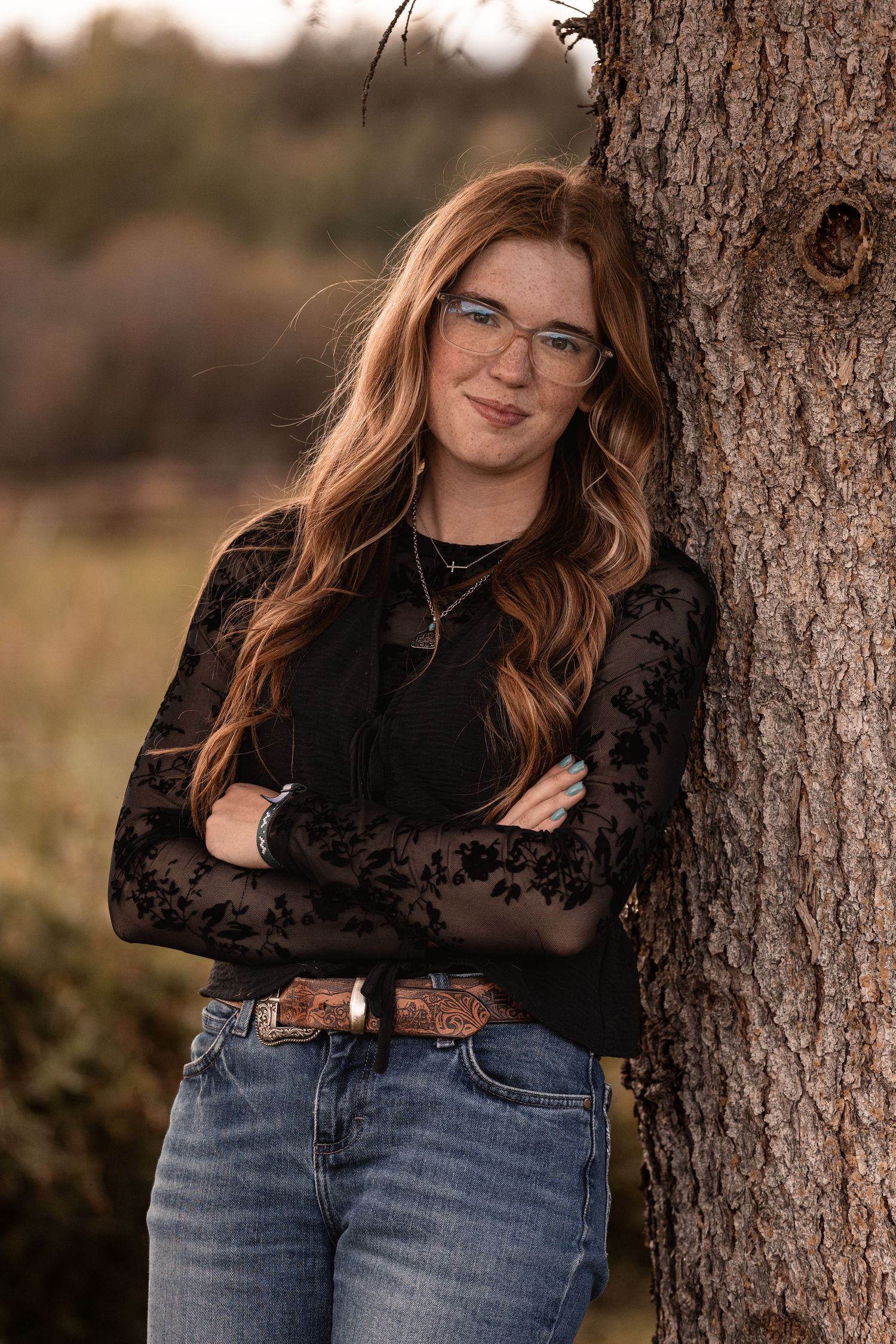 Young woman with red hair and glasses leans against a tree in jeans and black lace top outdoors.