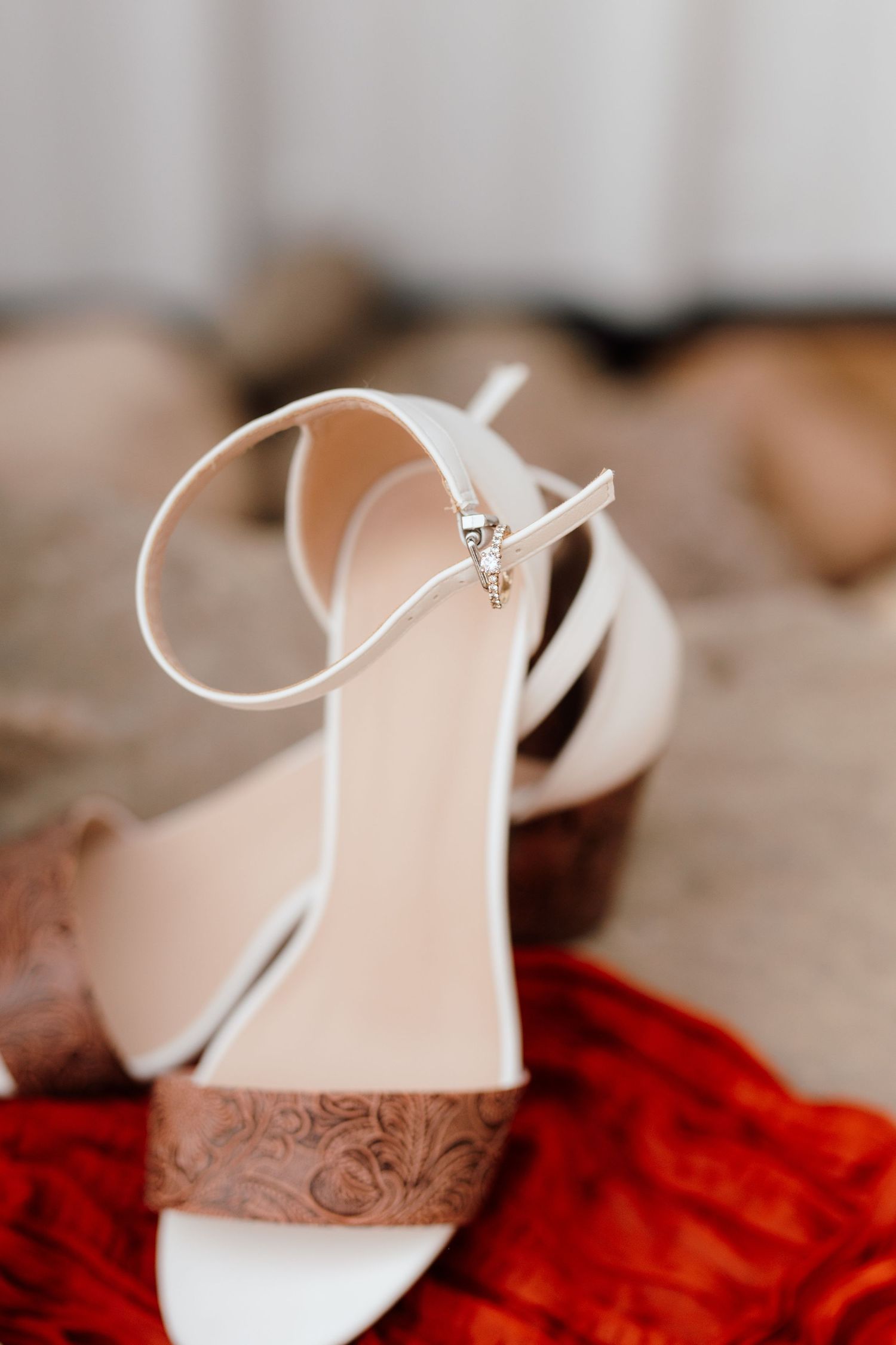 White bridal heeled sandals with ankle strap and ornate wood block heel resting on red fabric.