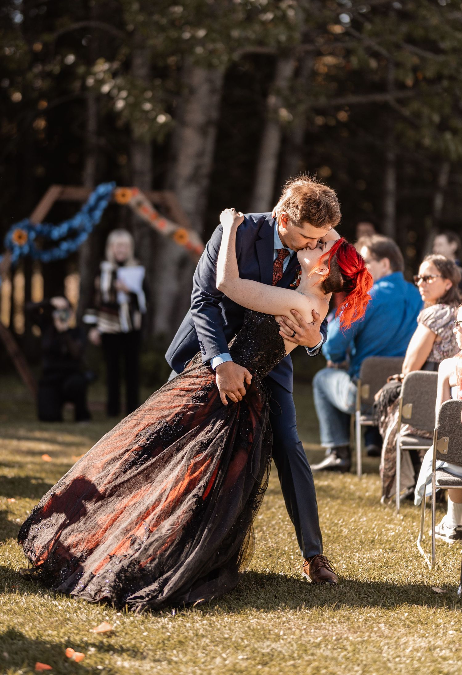 Couple sharing a romantic dip kiss at outdoor wedding ceremony, bride in black gown with red hair, groom in navy suit.