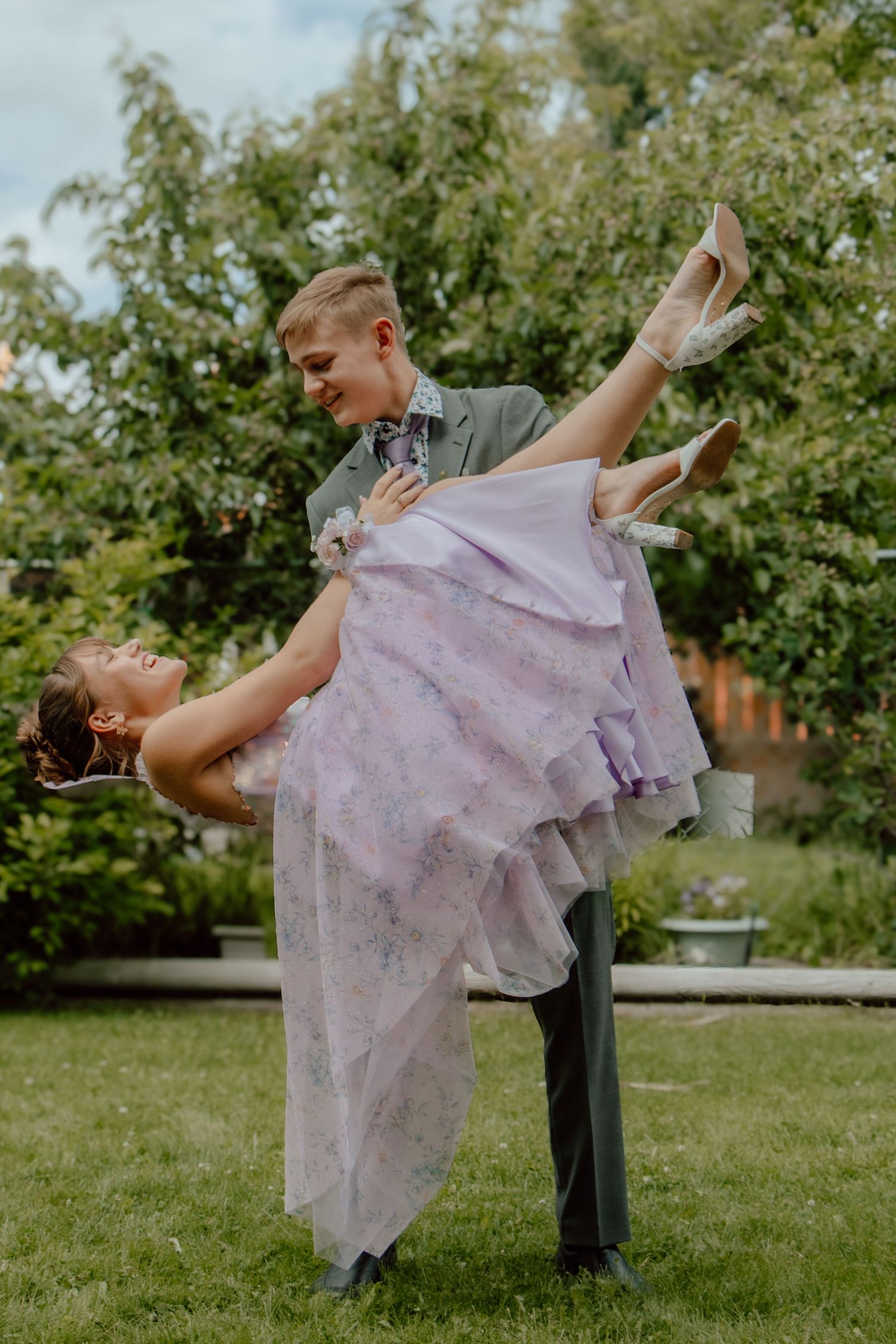 A joyful couple poses at prom, with the young man lifting the woman in a lavender floral dress.