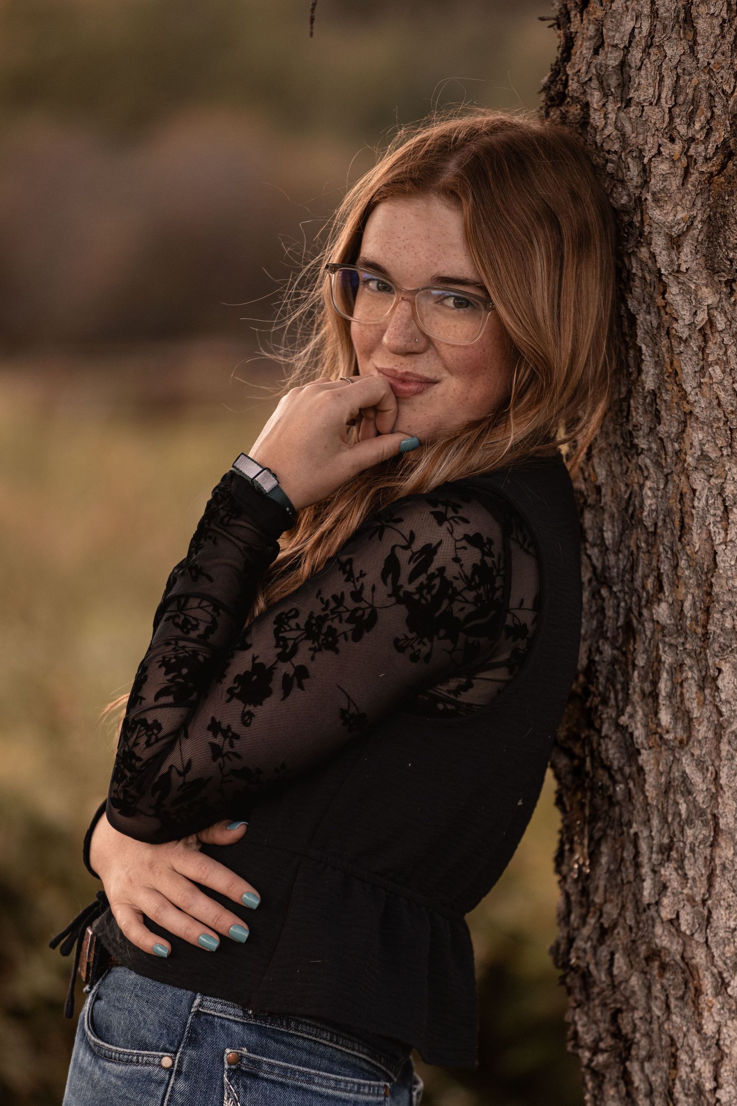 Young woman with glasses and red hair in black lace top leans against tree trunk outdoors.