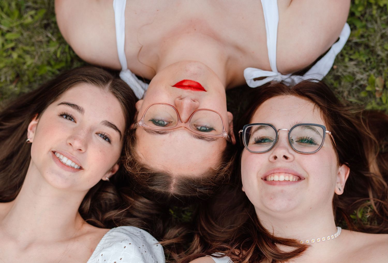 Three young women lying on grass, smiling and posing for a fun outdoor photo together.