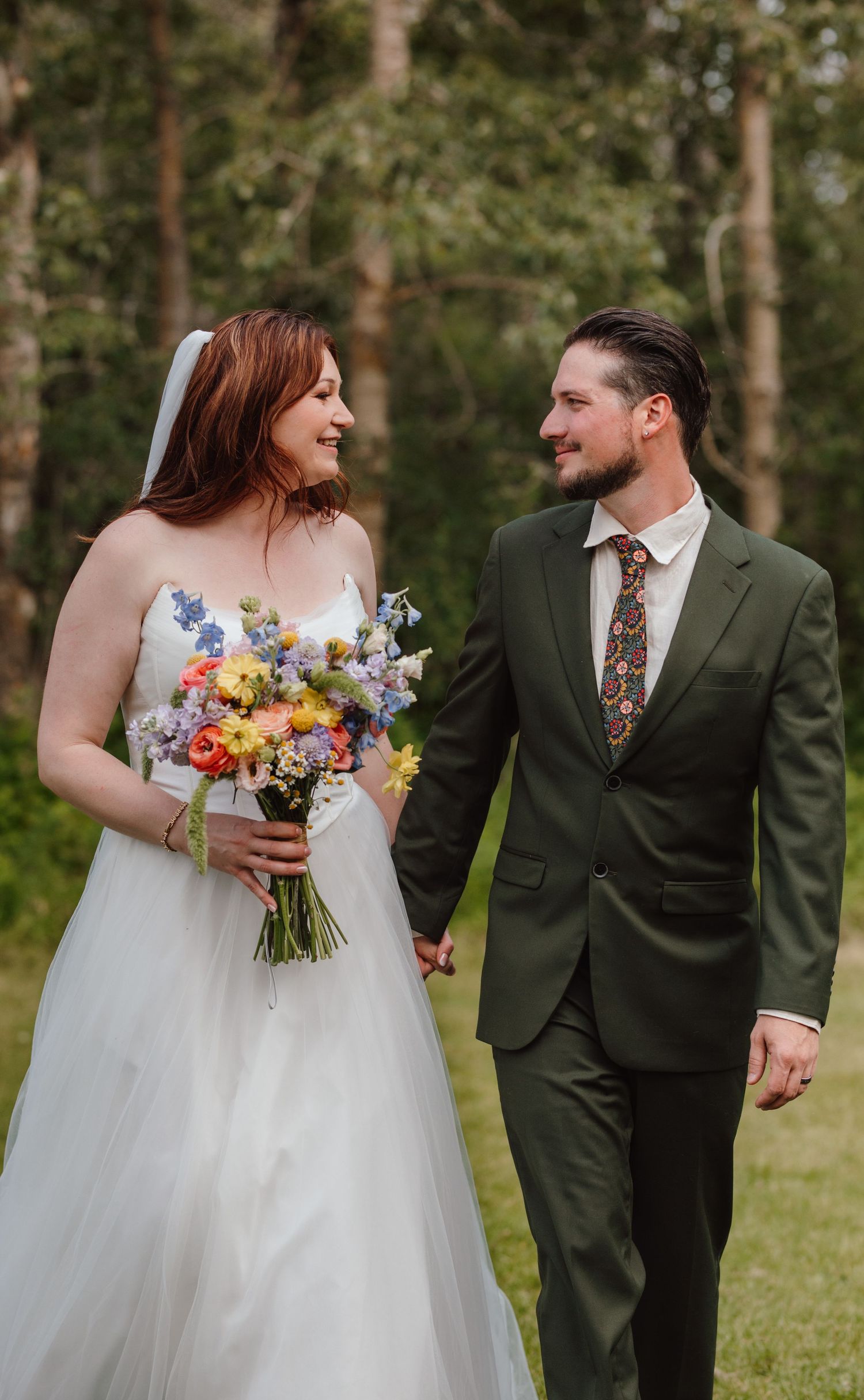 Bride and groom walking hand in hand outdoors, she holds a colorful wildflower bouquet, both smiling at each other.