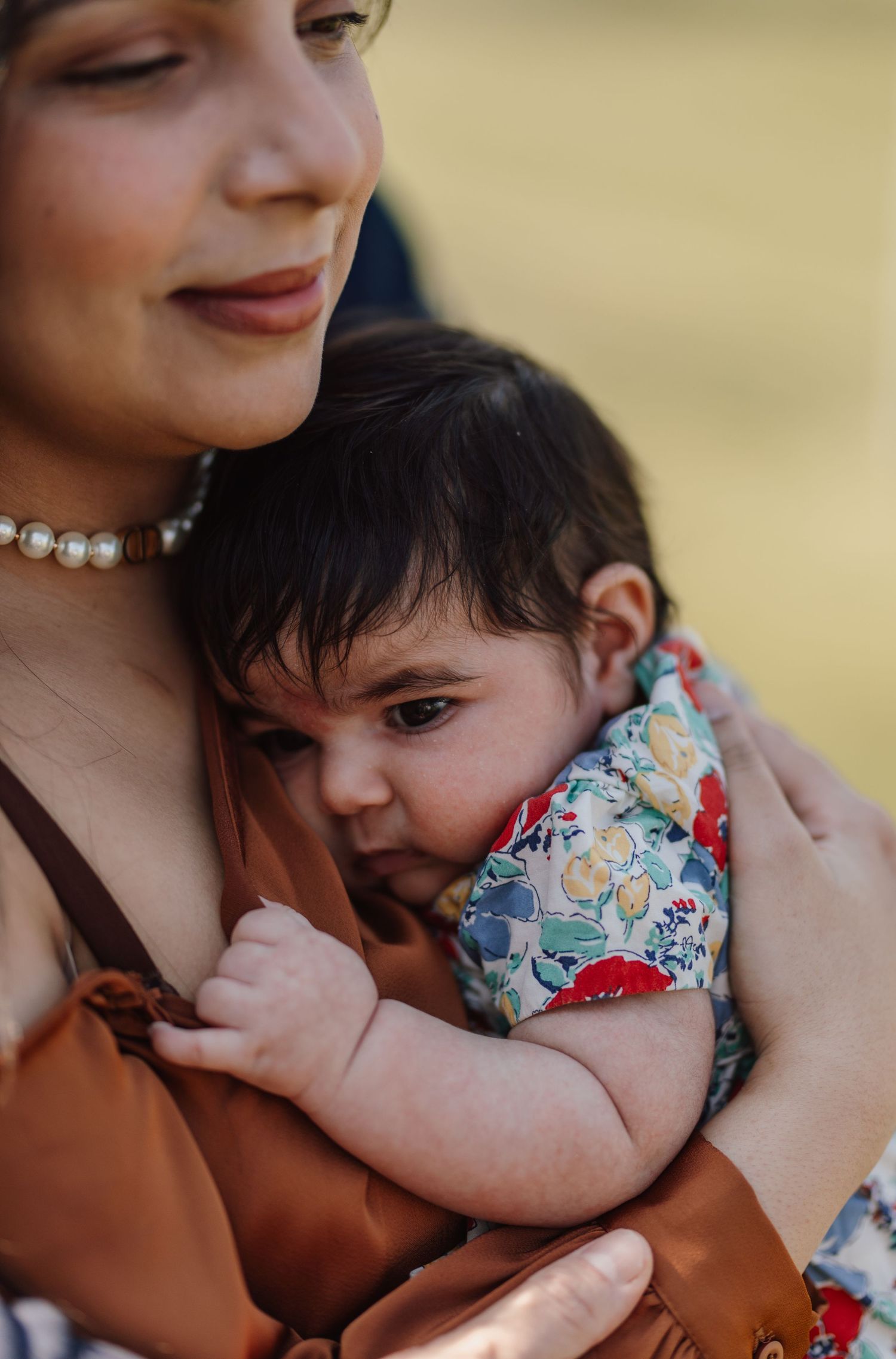 A mother wearing a pearl necklace tenderly holds her dark-haired baby dressed in a colorful floral outfit outdoors.