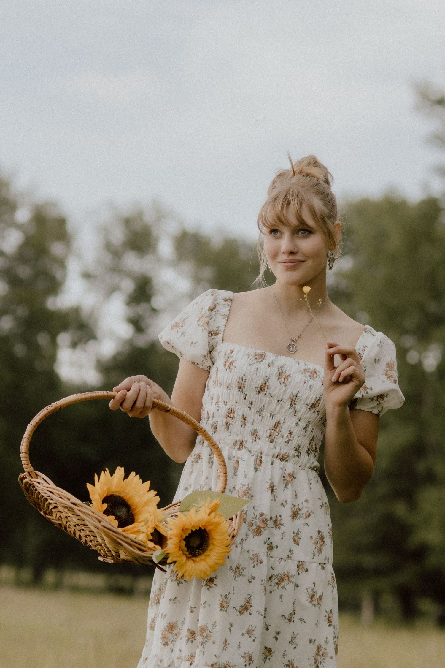 Young woman in white floral dress holding a wicker basket with sunflowers in a scenic outdoor park setting.