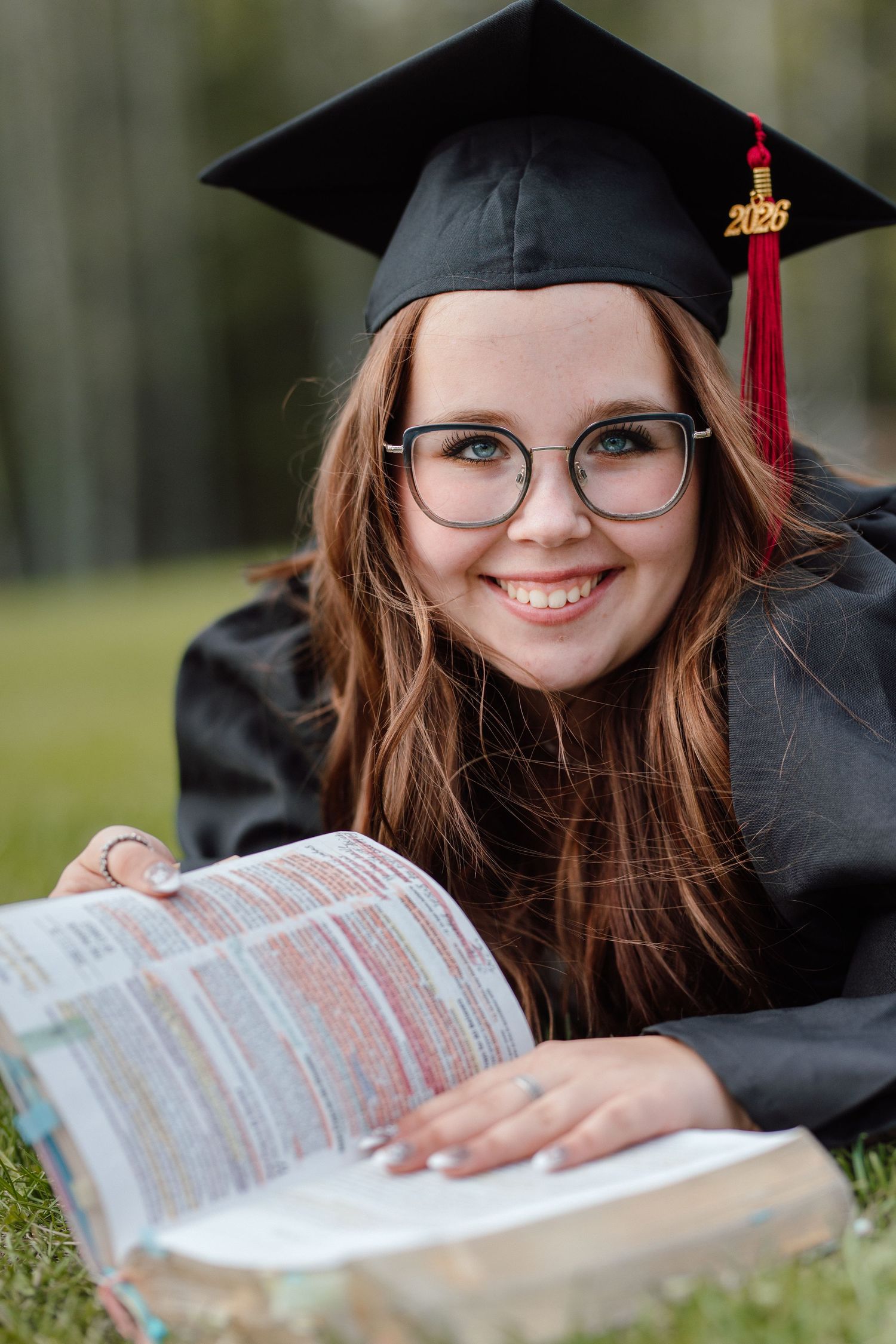 Smiling graduate girl in cap and gown lying on grass reading a highlighted Bible outdoors in a park setting.
