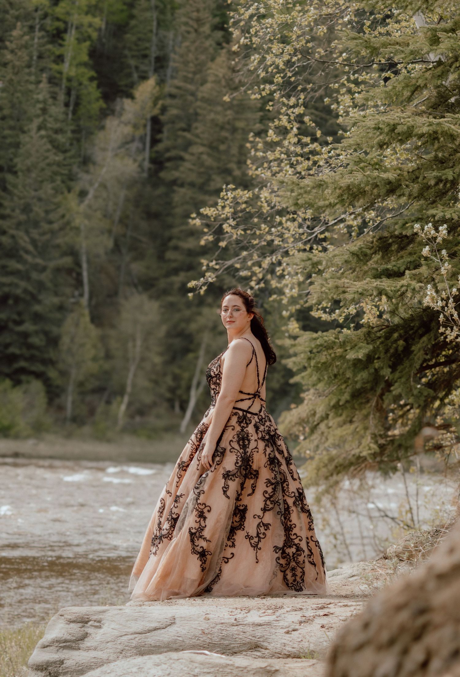 A woman in a stunning rose gold and black lace ball gown poses gracefully by a mountain river.