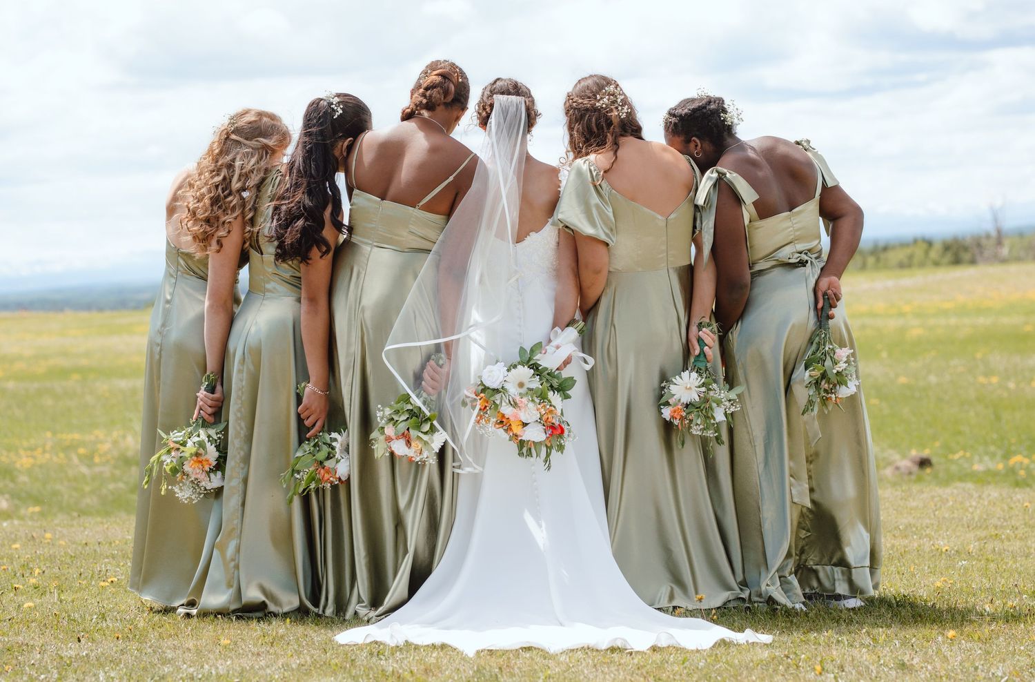 Bride and bridesmaids in sage green gowns holding bouquets, posing with backs to camera in a field.