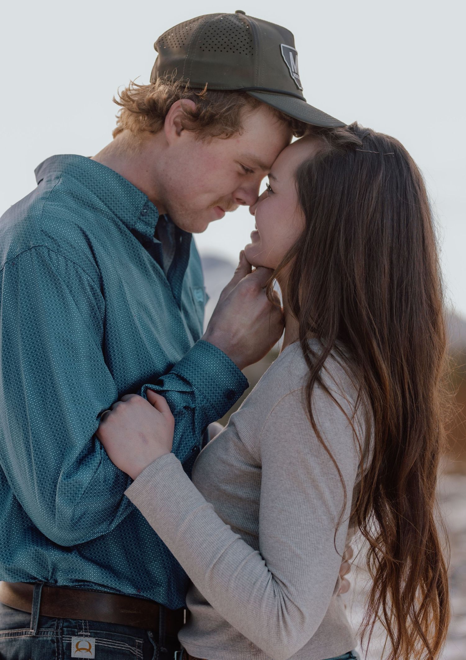 A young couple shares a romantic forehead touch outdoors, the man in a green cap and blue shirt embracing a brunette woman.