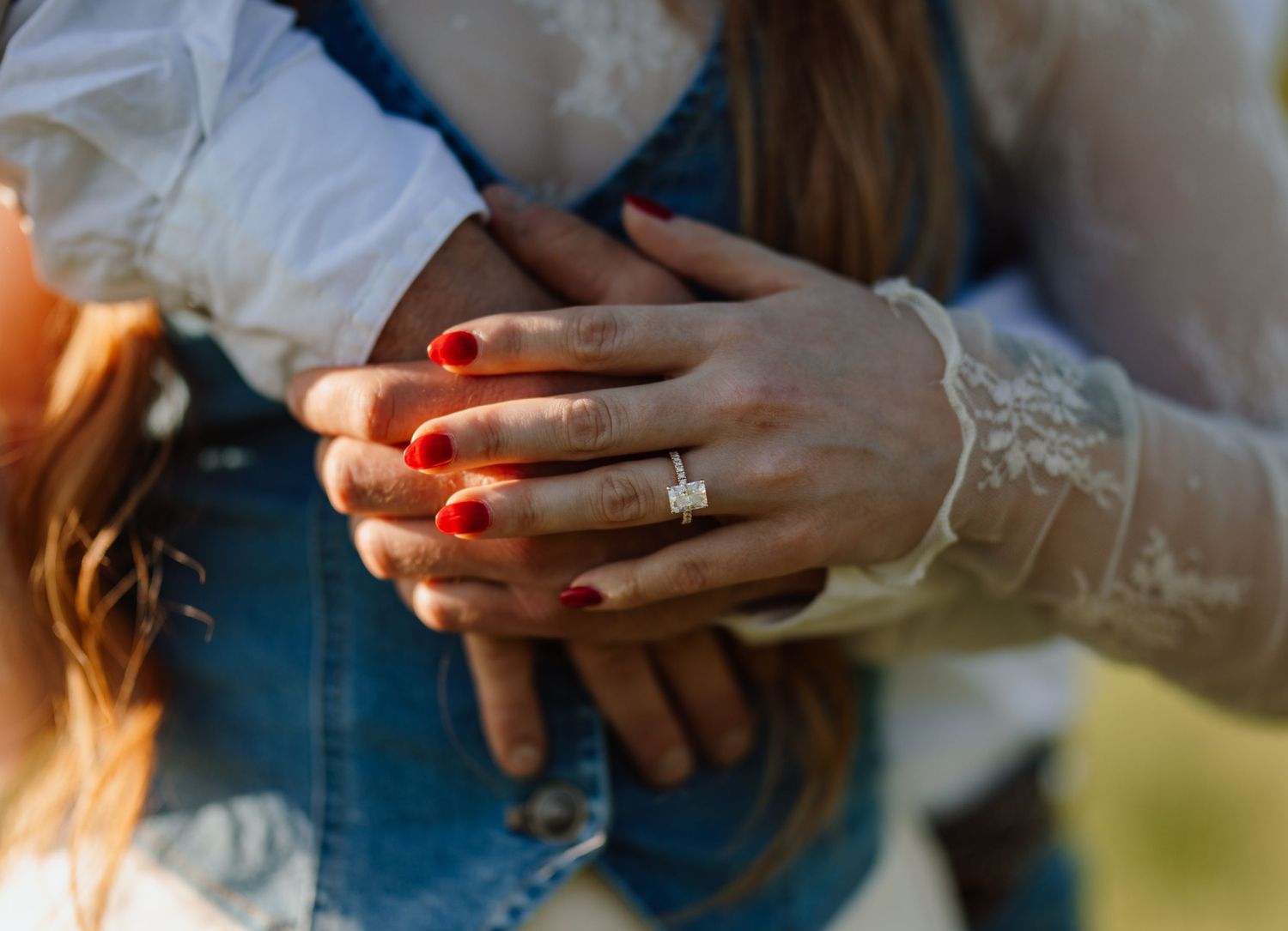 Close-up of woman's hands with red nails and diamond engagement ring, embraced from behind by partner.