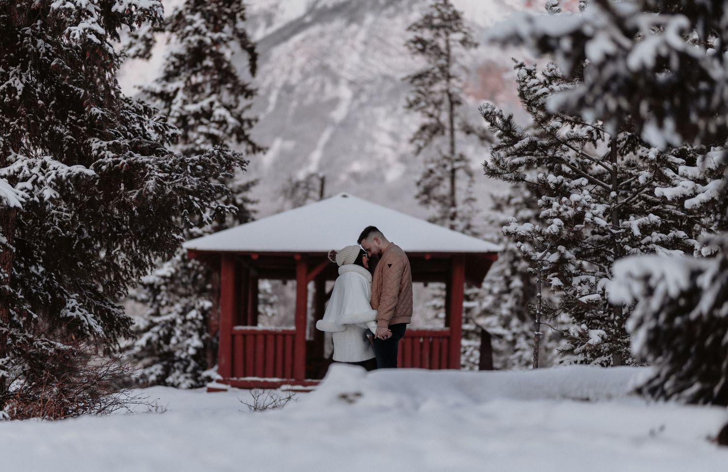Couple embracing in snow near red gazebo surrounded by snow-covered pine trees and mountains.