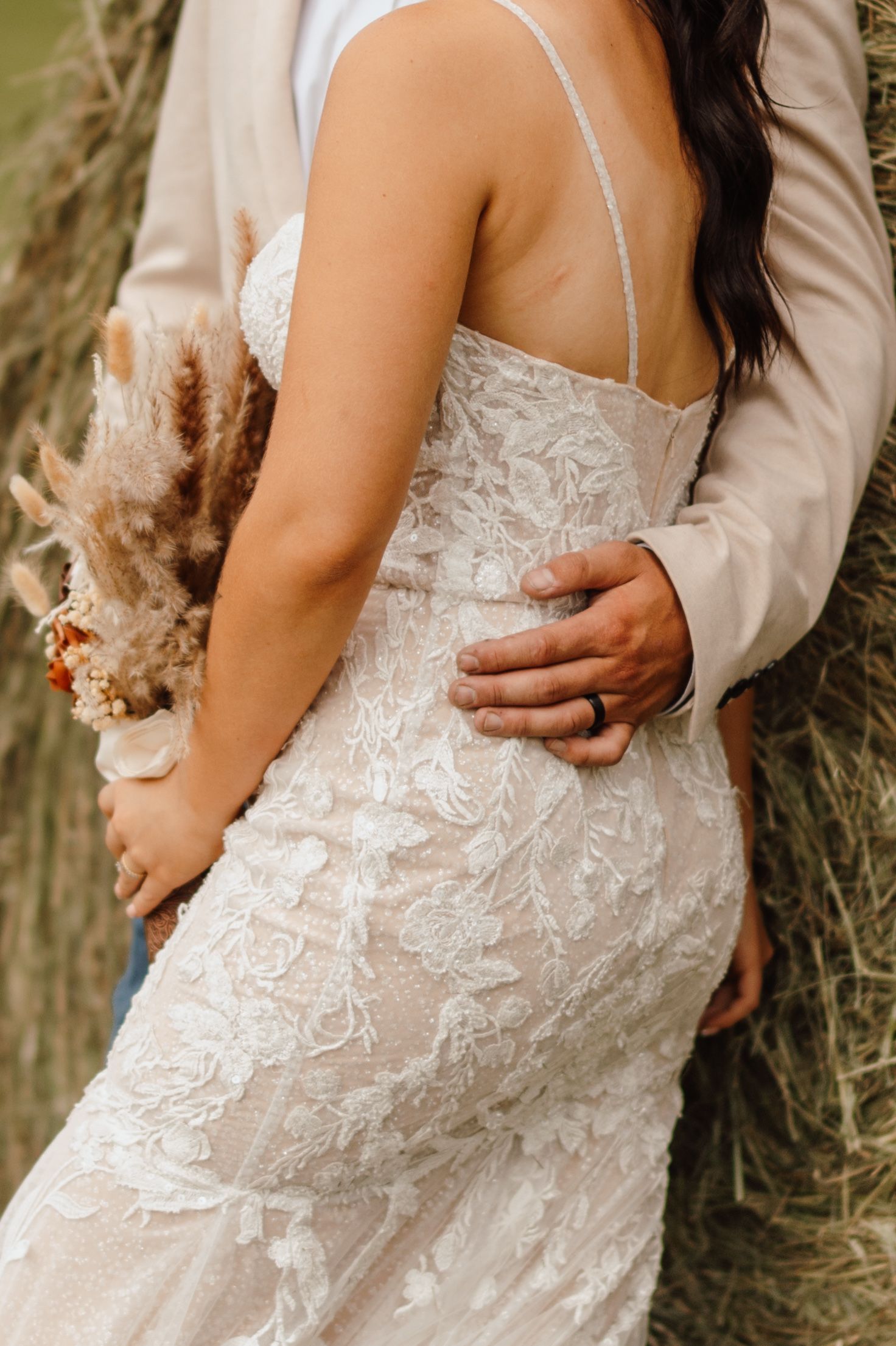 A bride in a lace wedding gown embraced from behind by a groom in a beige suit, holding a dried floral bouquet.