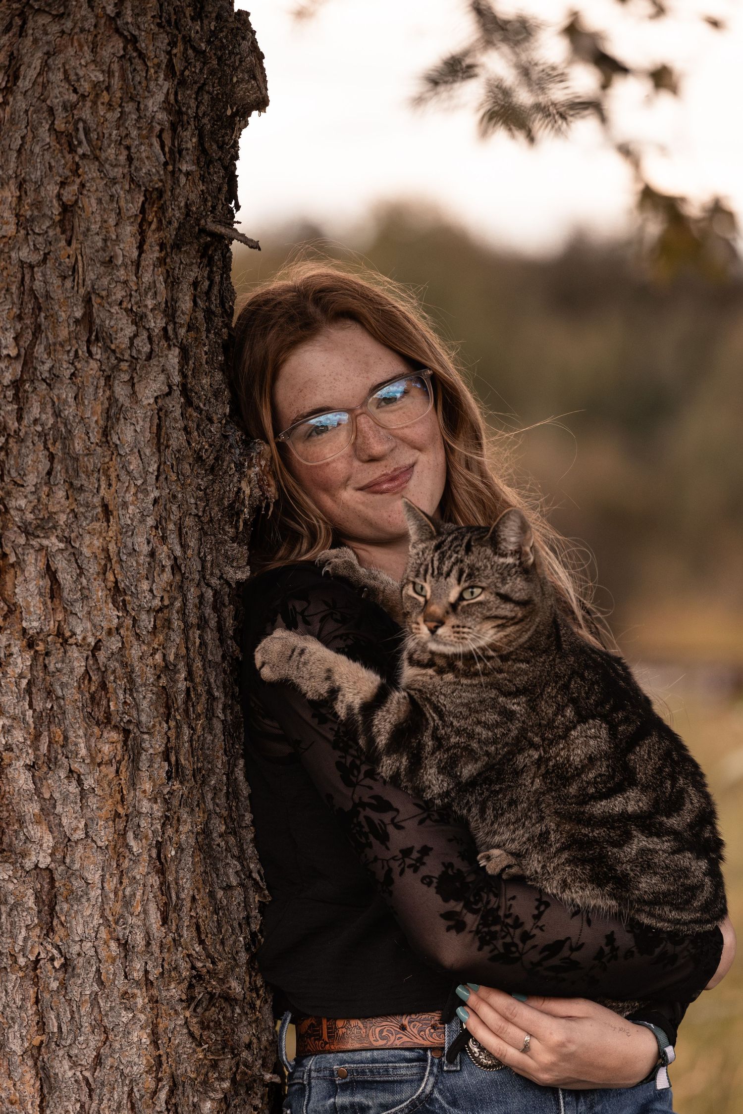 A smiling redhead woman with glasses holds a tabby cat while leaning against a tree outdoors.