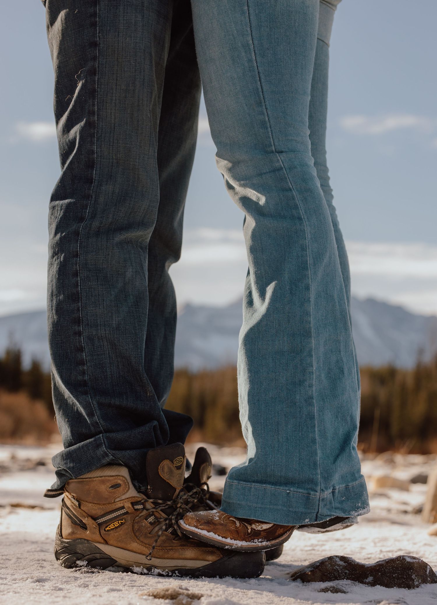 Close-up of two people in jeans and hiking boots standing in snow with mountains in the background.