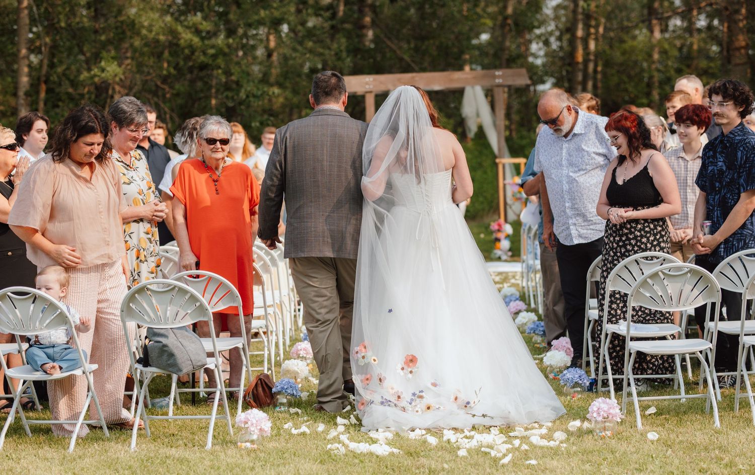 Bride in white gown with floral veil walks down outdoor aisle with guests seated on white chairs.
