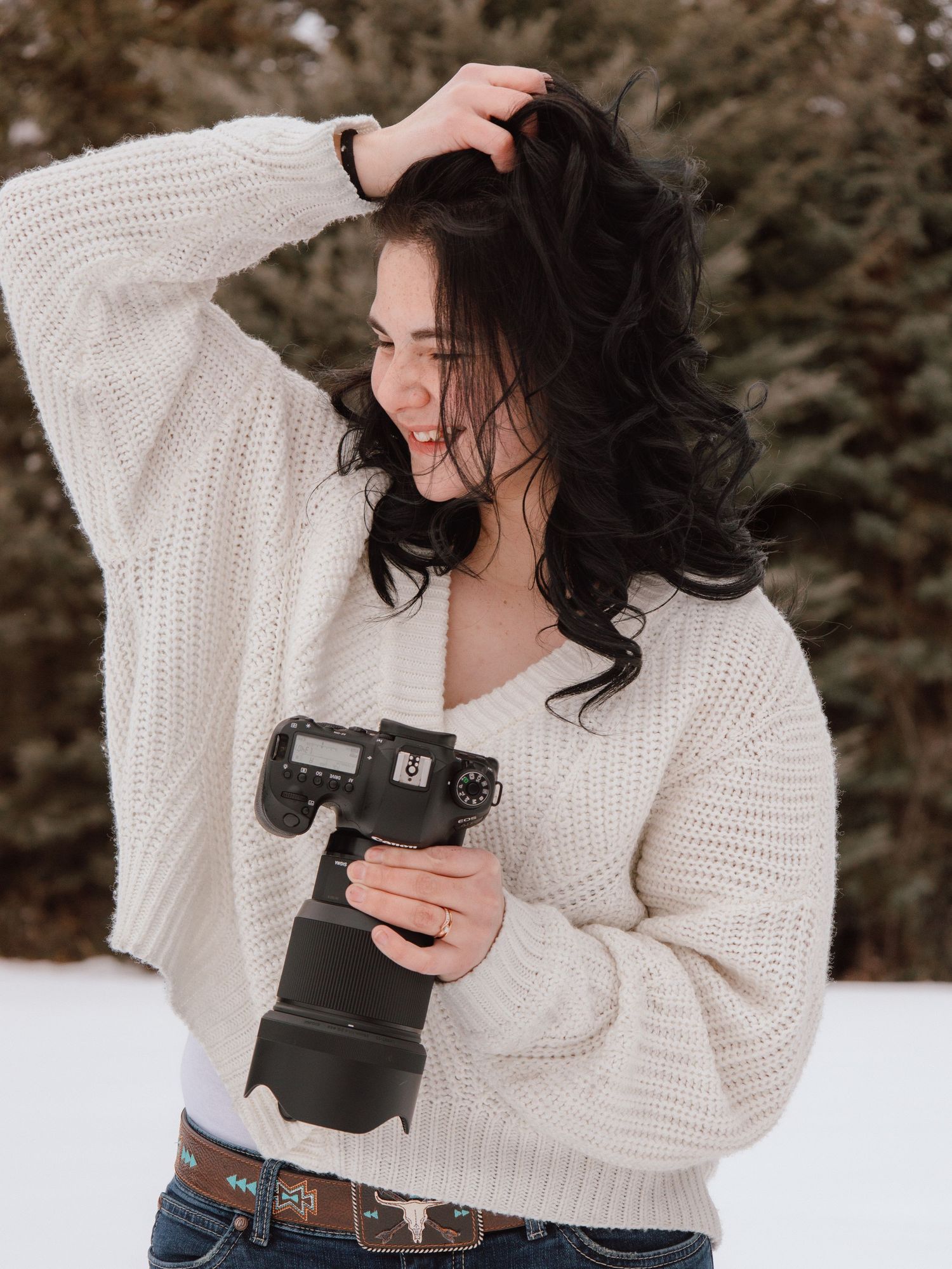 A young woman in a white knit sweater holds a DSLR camera while posing outdoors in a snowy winter setting.