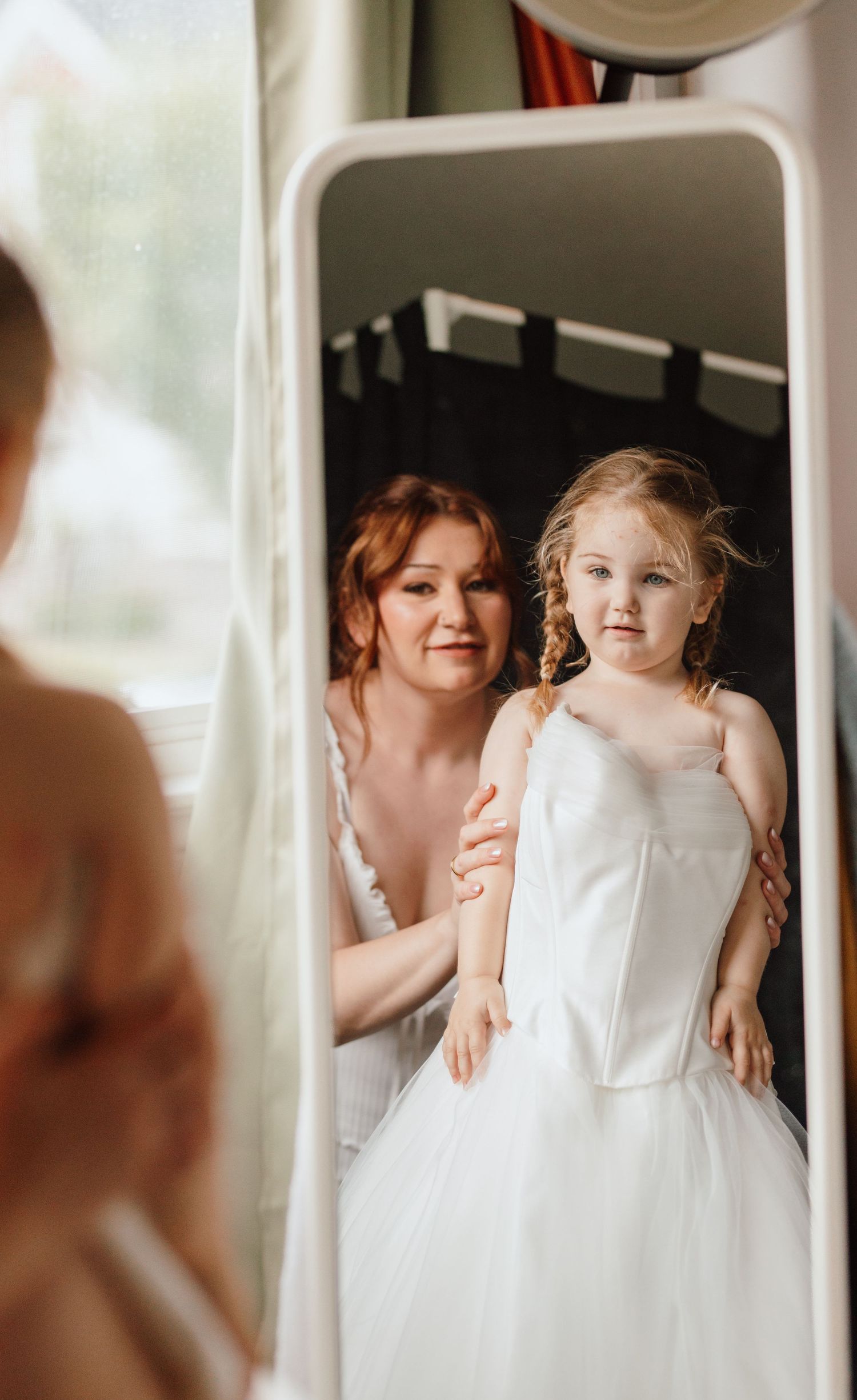 A bride in a white gown kneels behind a young flower girl in a white dress, both reflected in a mirror.