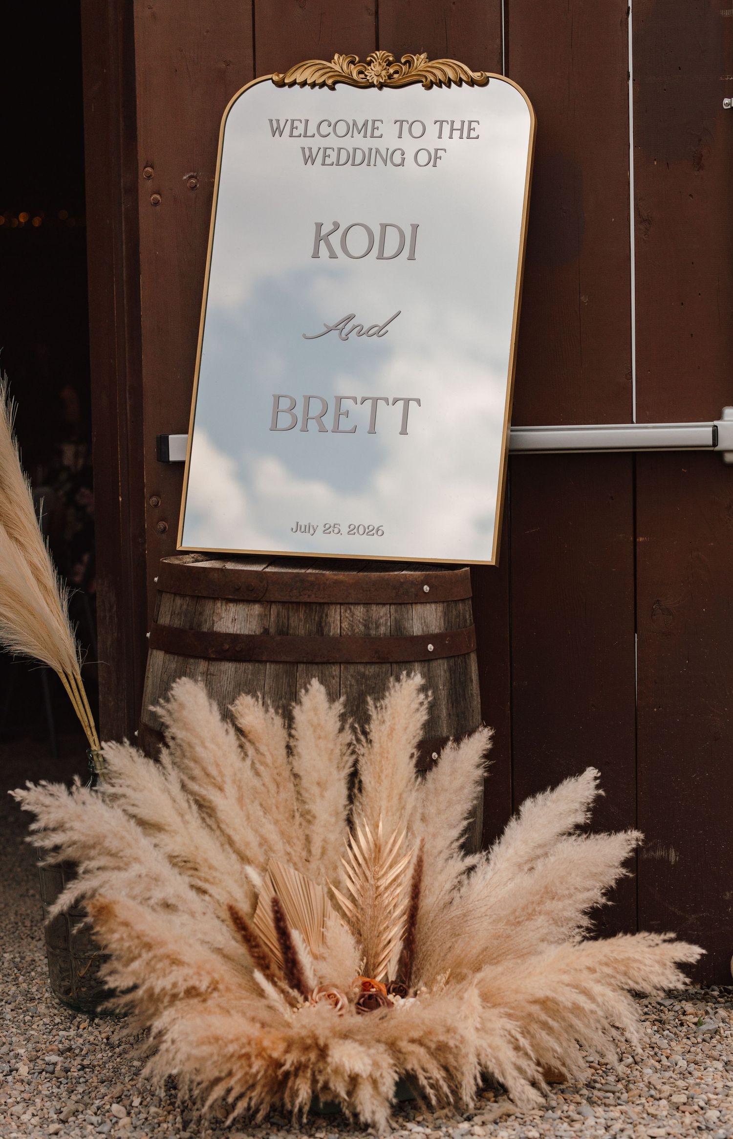 Wedding welcome mirror sign for Kodi and Brett on a rustic barrel, adorned with pampas grass decor.