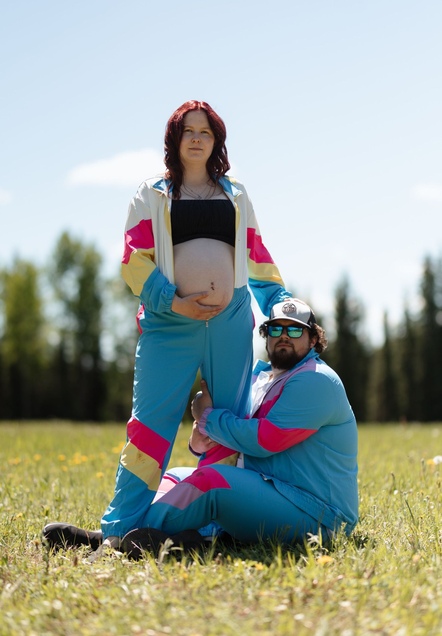 Pregnant woman and man in matching retro 80s blue tracksuit outfits posing outdoors in a sunny green field.