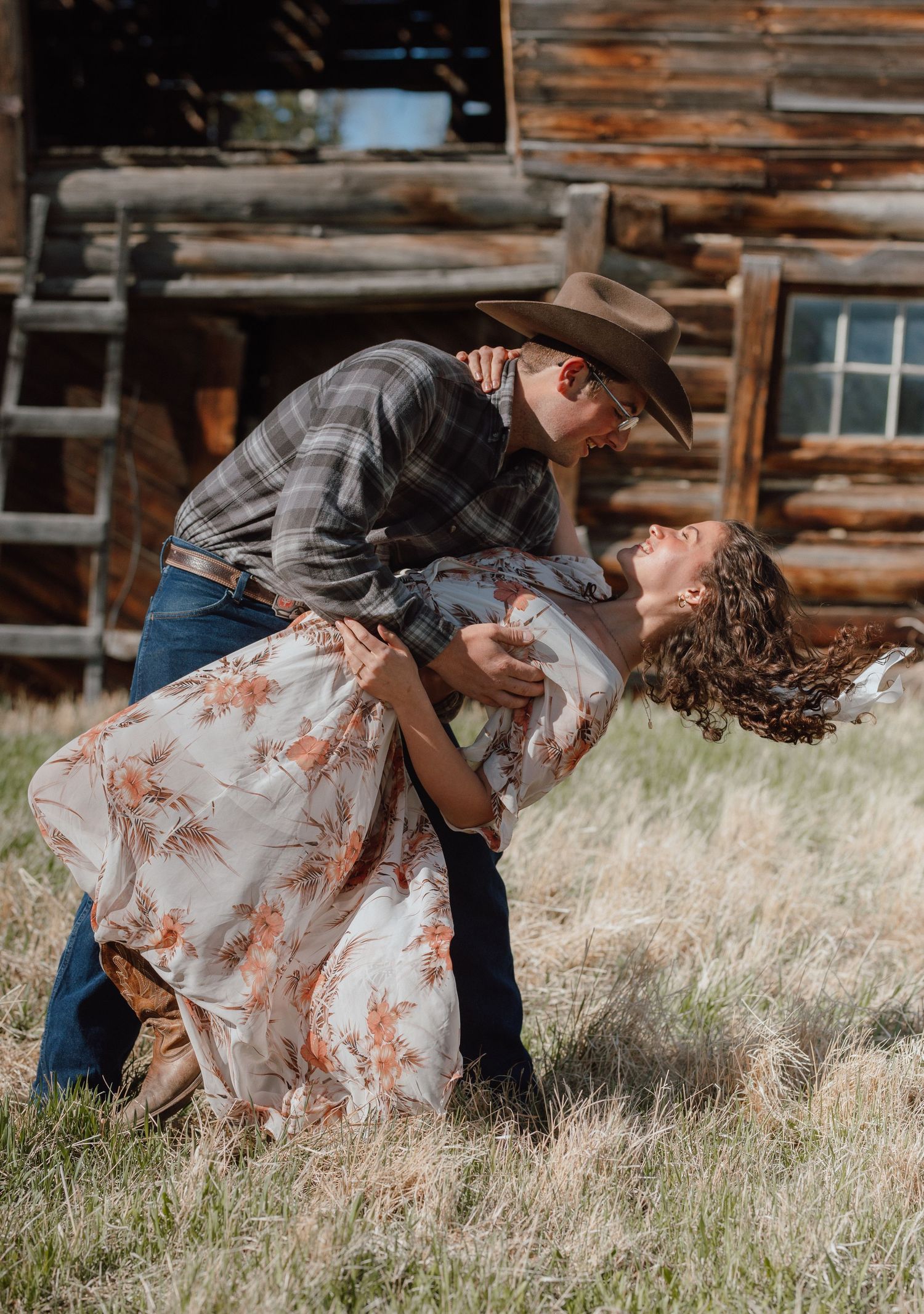 Couple sharing romantic dip kiss in field near rustic log cabin in western attire.