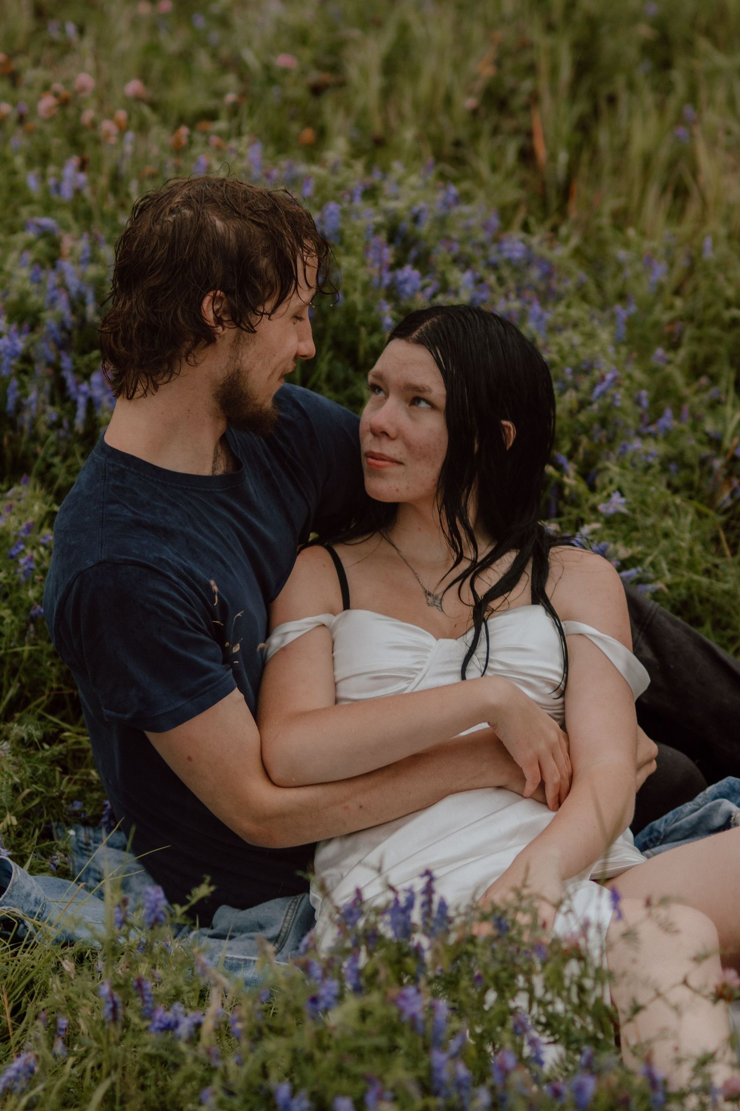 A couple sits intimately together in a field of purple wildflowers, gazing into each other's eyes.