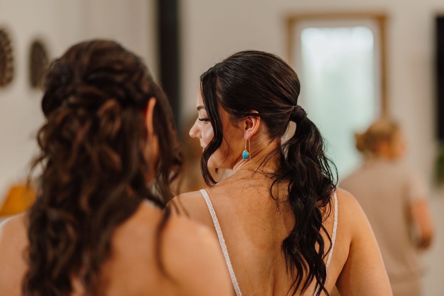 Two bridesmaids with elegant updo hairstyles and turquoise earrings prepare together before a wedding ceremony.