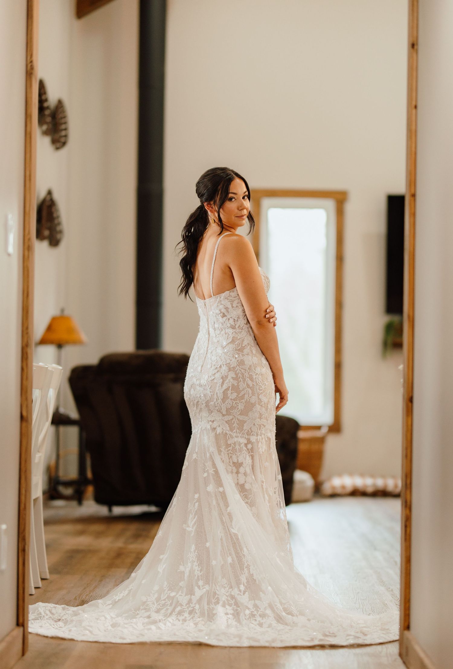 Bride in elegant lace mermaid wedding gown with train, posing in doorway looking over shoulder.
