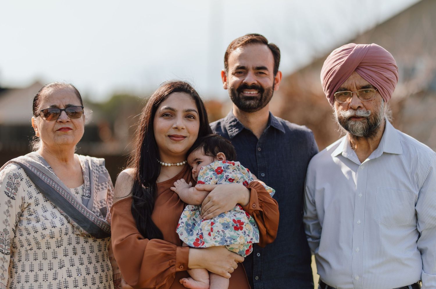 A Sikh family of four poses outdoors, with parents holding a baby, grandparents beside them, smiling warmly.
