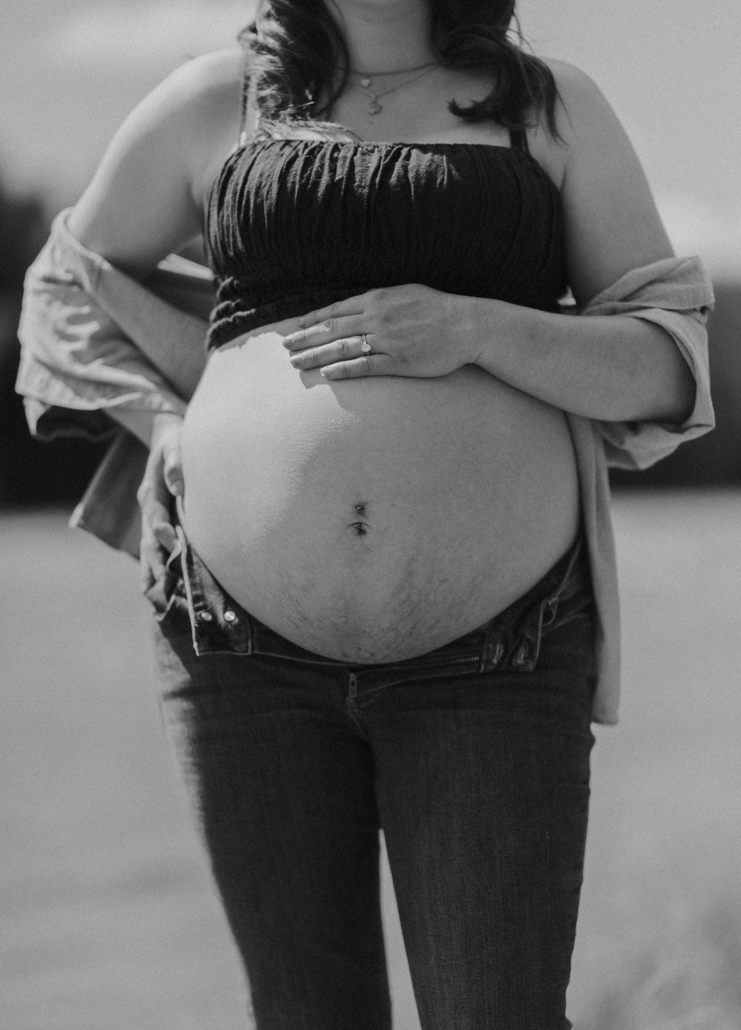 Black and white maternity photo of pregnant woman cradling bare baby bump, wearing crop top and jeans outdoors.