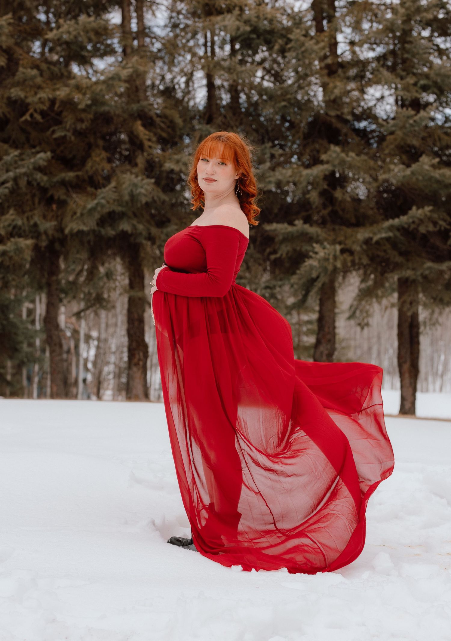 Pregnant woman in flowing red off-shoulder gown standing gracefully in a snowy forest setting.