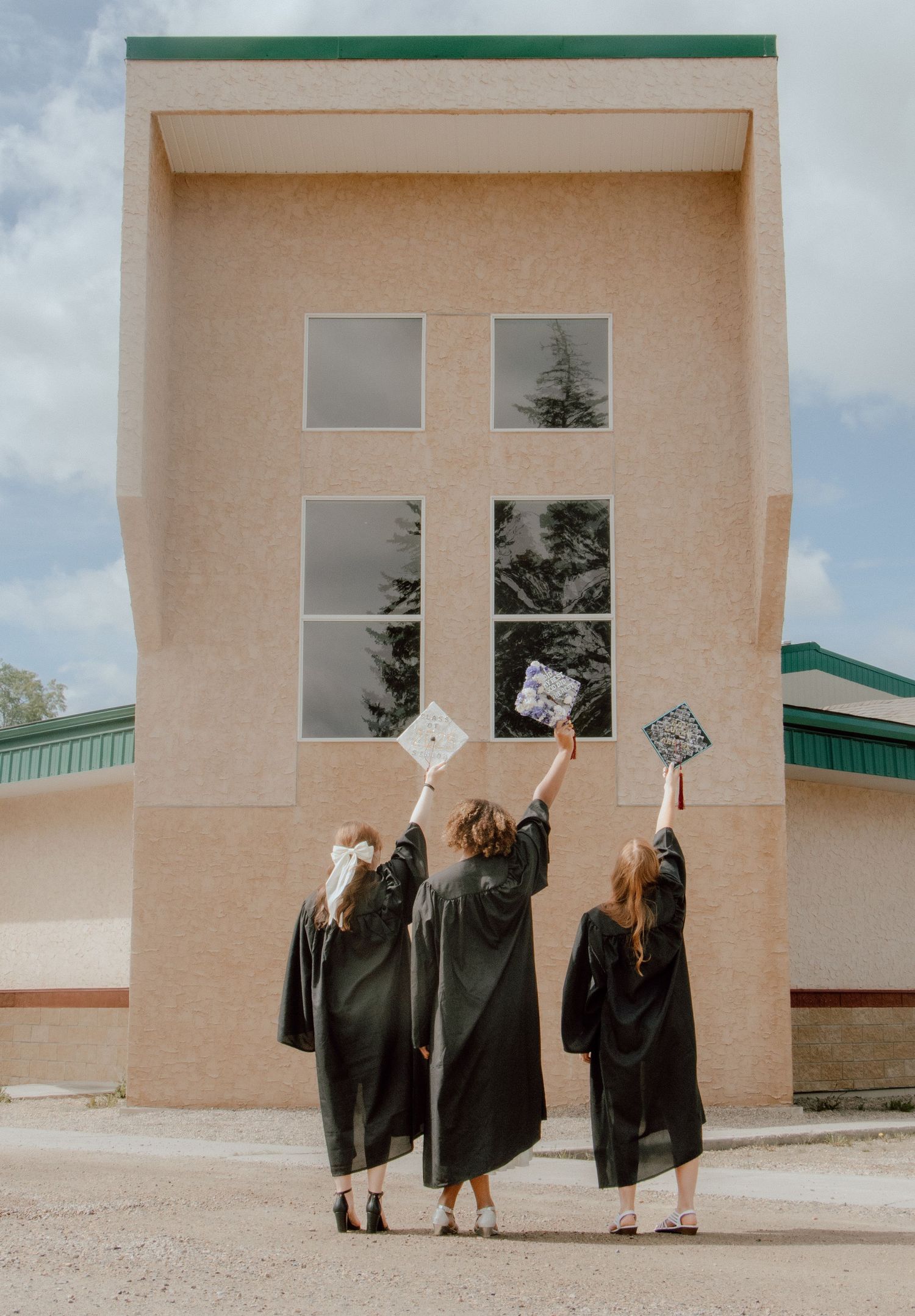Three graduates in black gowns raise their decorated caps toward a building, celebrating their academic achievement outdoors.
