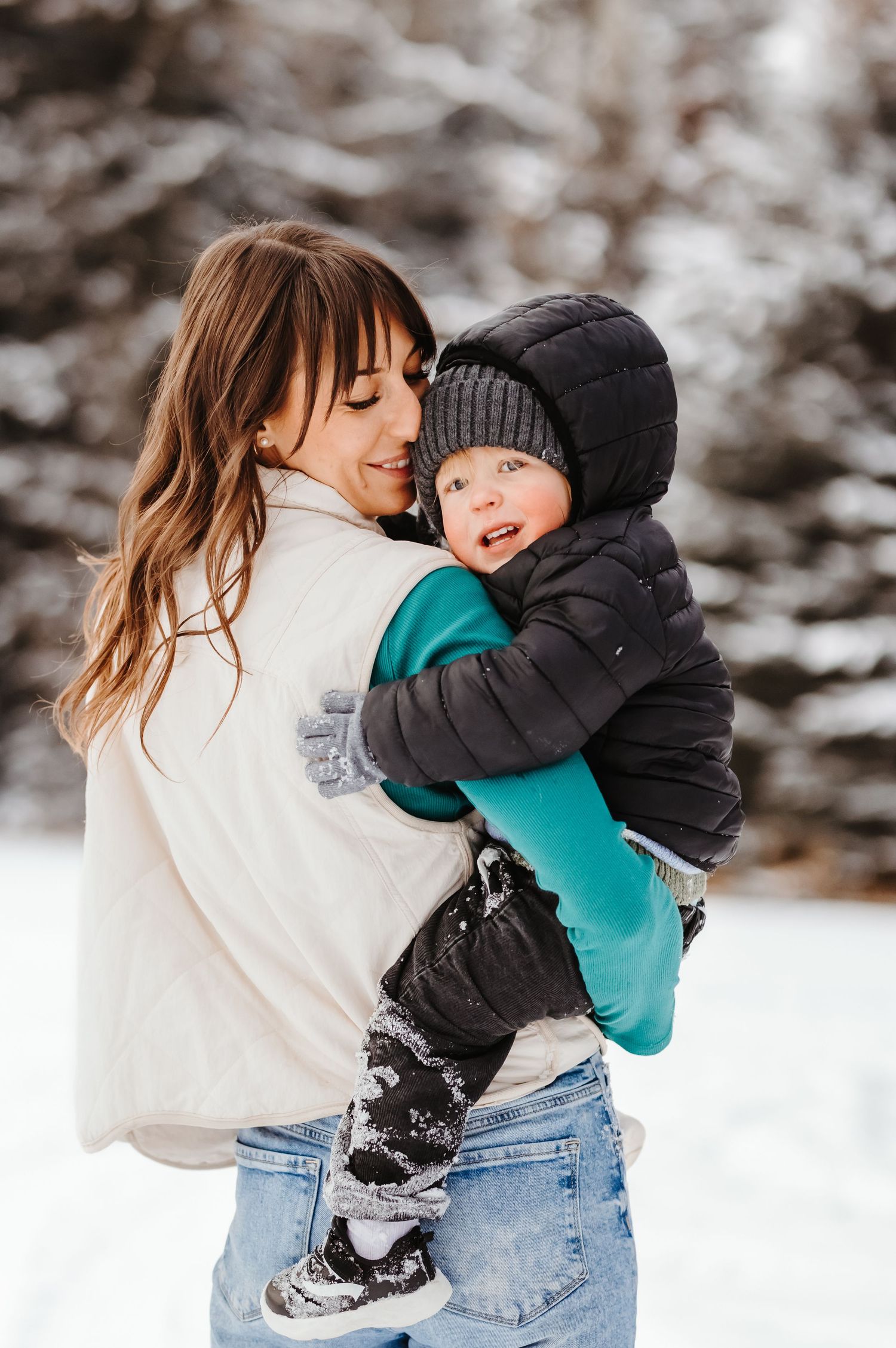 Smiling mother in white vest holding bundled toddler in snowy winter forest landscape.