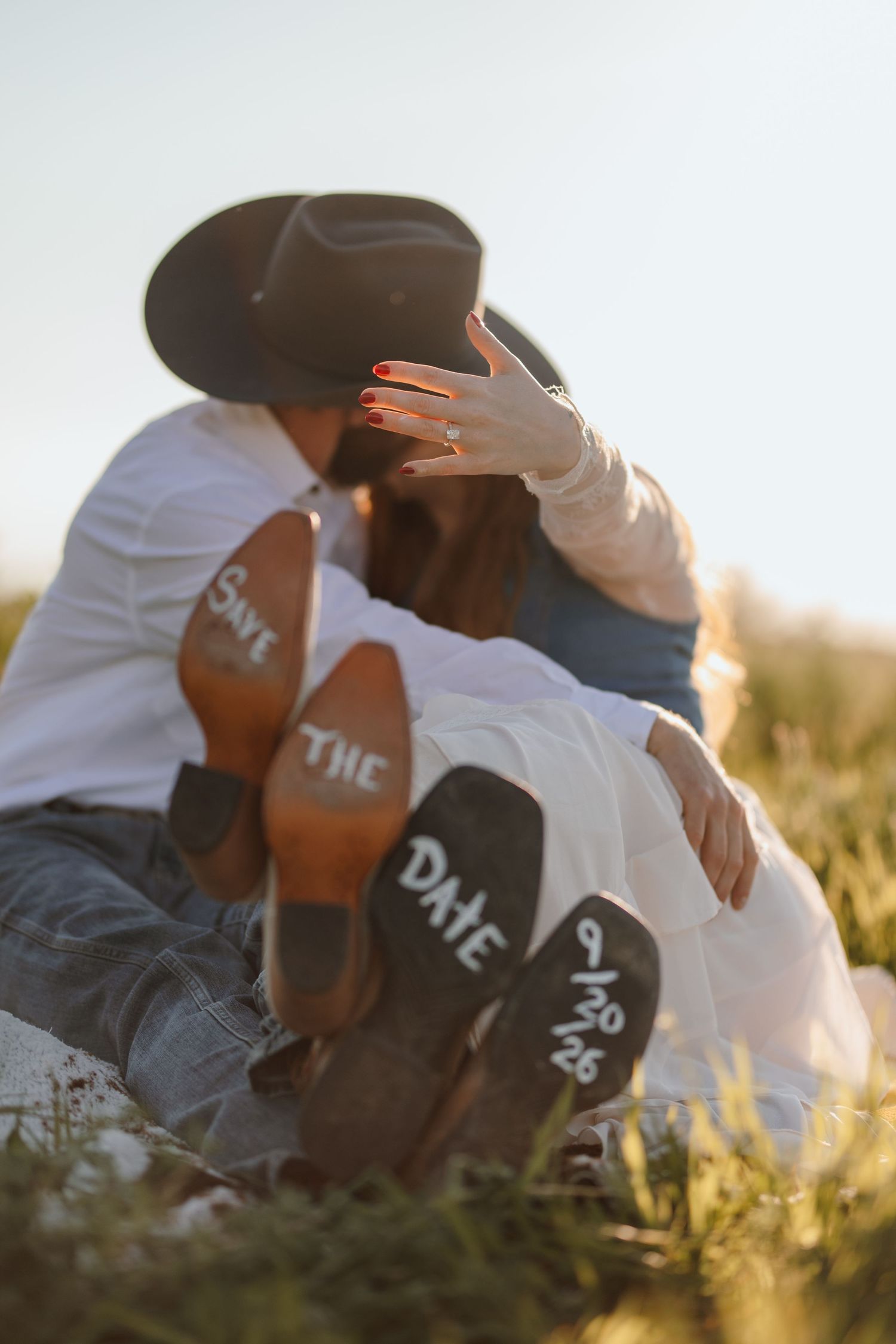 Couple sitting in field kissing with save the date 9/30/26 written on cowboy boot soles.