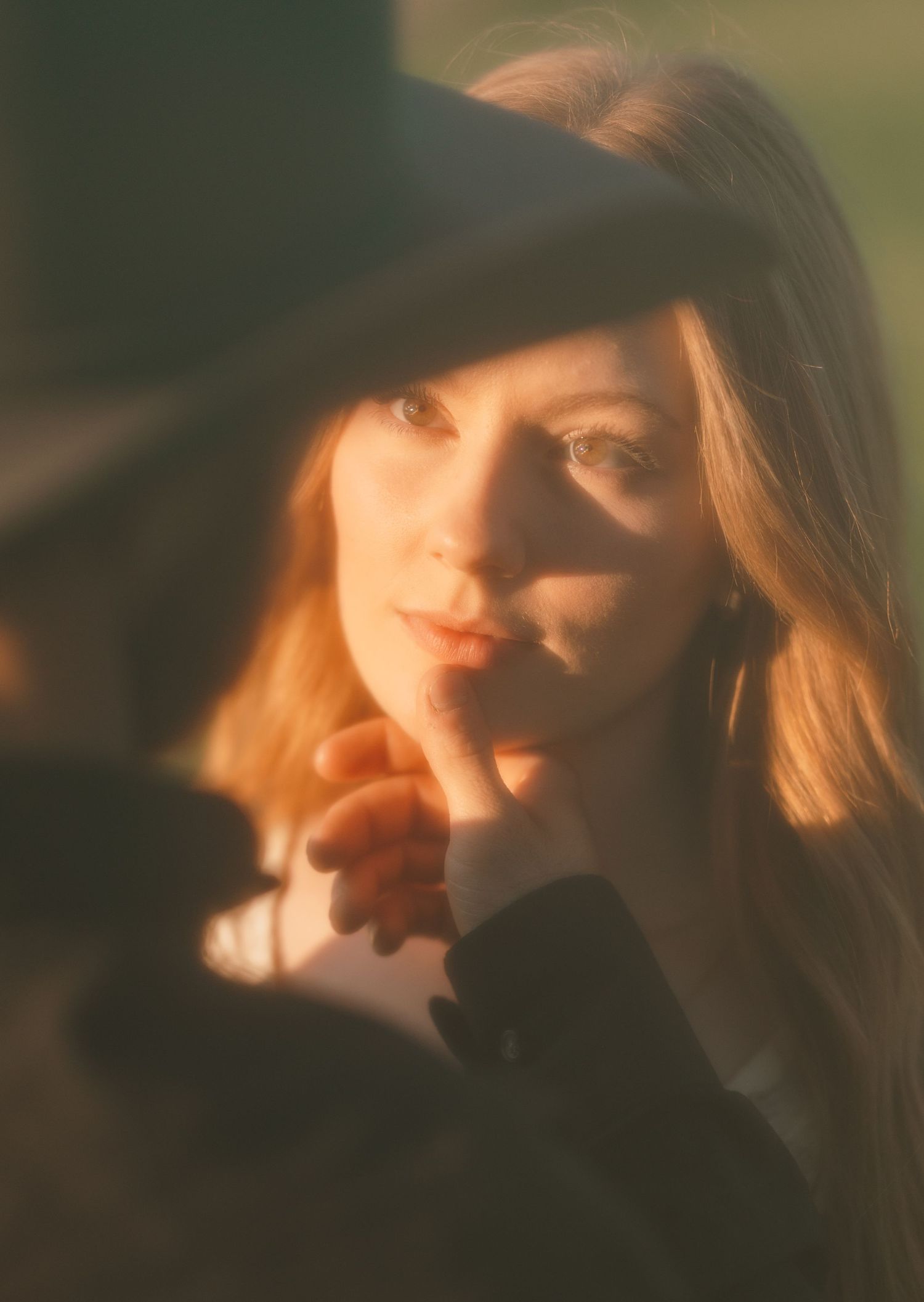 A woman with long auburn hair wearing a black hat, touching her lips in warm golden sunlight.
