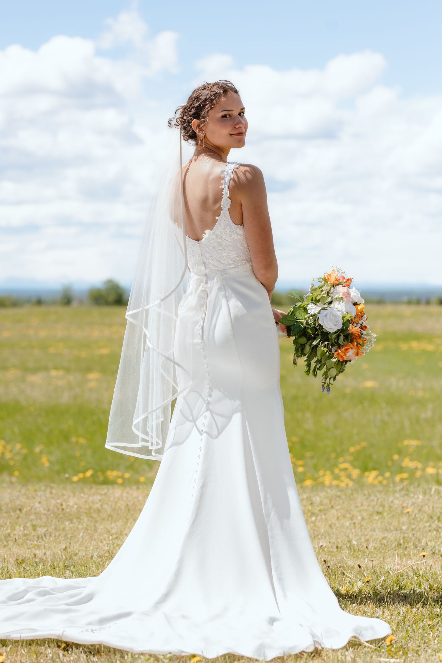 Bride in white backless gown with veil holds bouquet in sunny field.