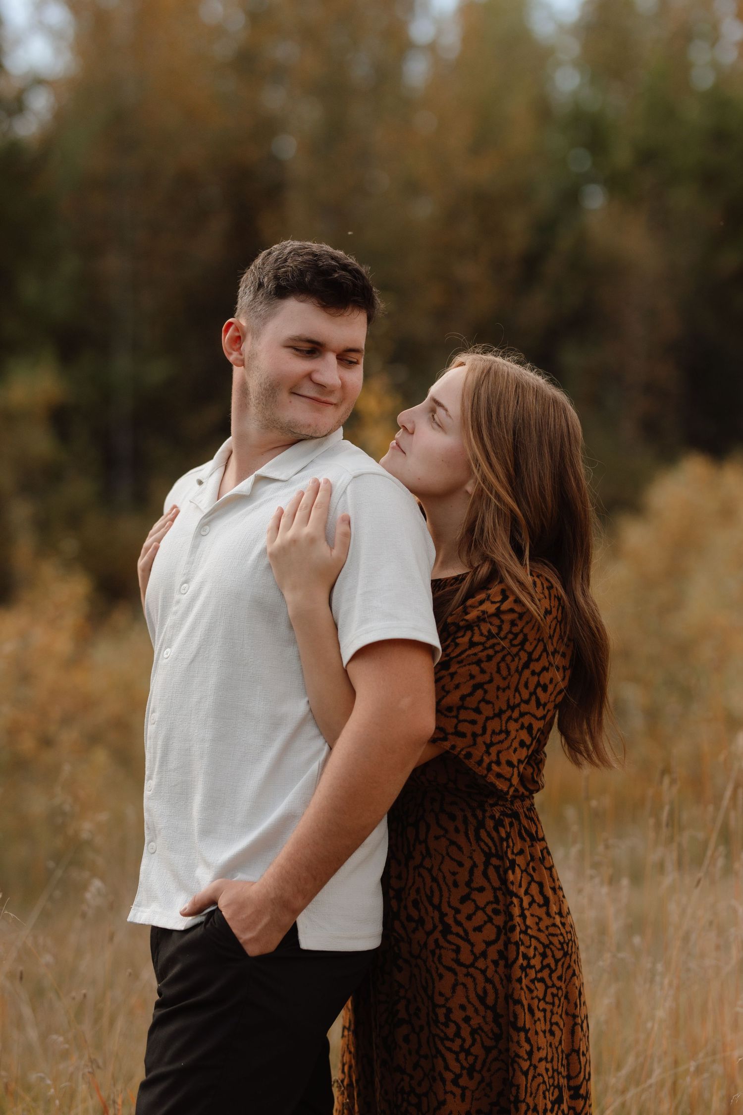 Couple embracing in autumn field, woman in leopard print dress leaning on mans shoulder in white shirt.
