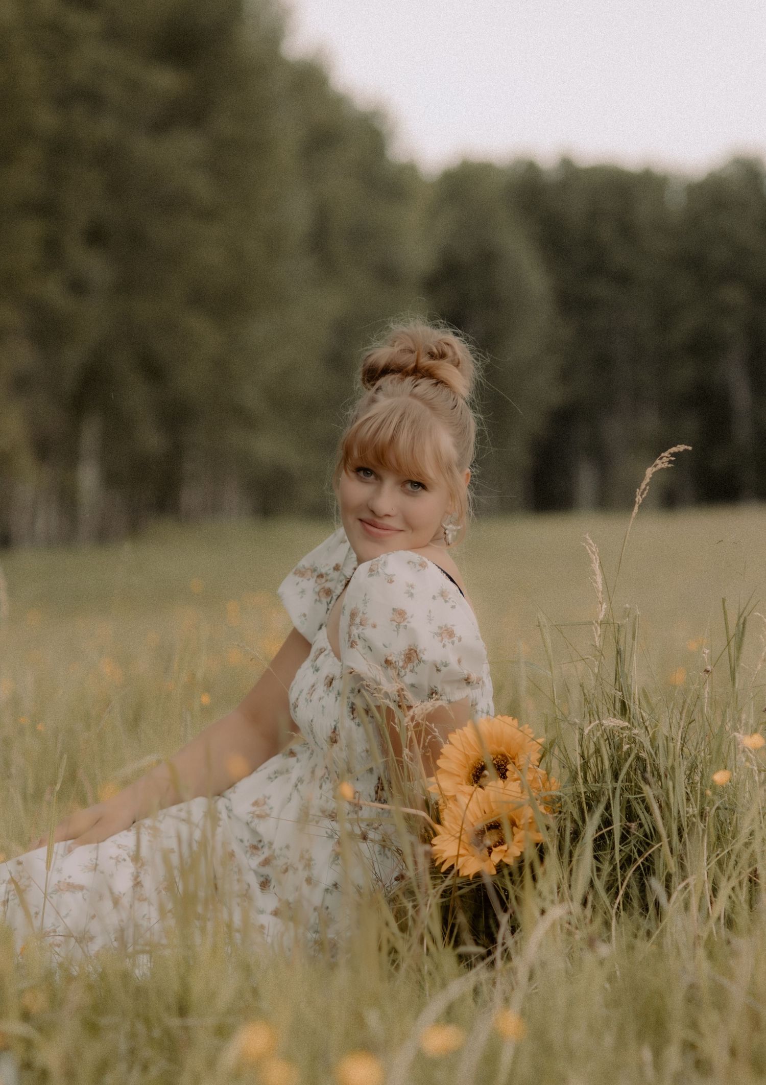 Young woman in floral dress sitting in a meadow holding sunflowers, smiling over her shoulder.
