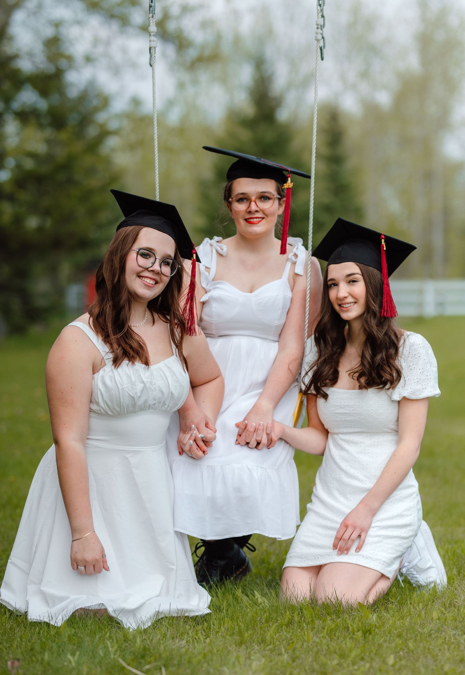 Three female graduates in white dresses and black caps with red tassels posing together on a swing outdoors.