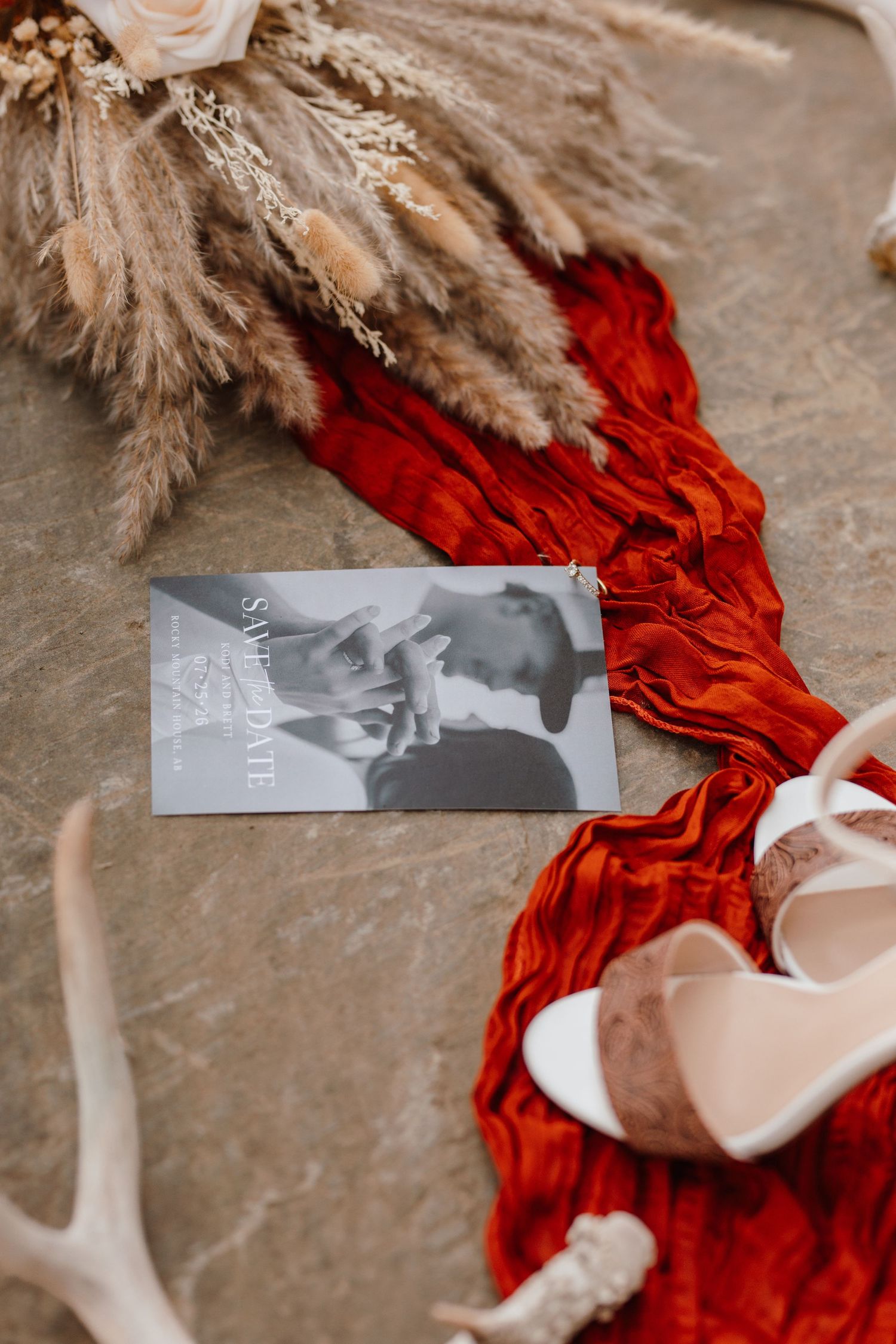 Save the date card with couple photo on red fabric beside white heels, pampas grass, and antlers on stone surface.