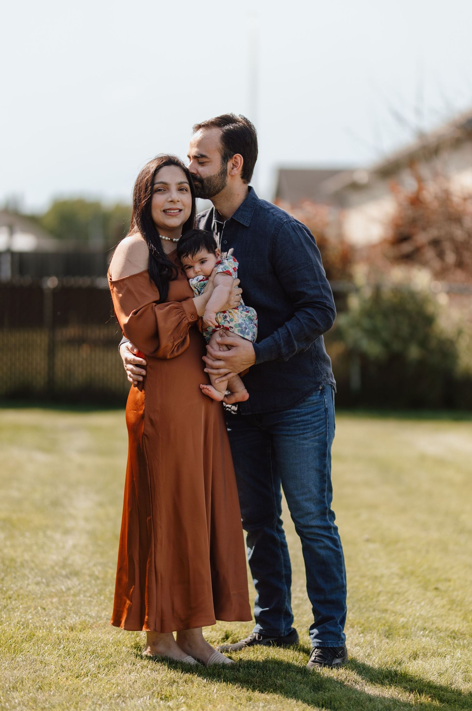 A happy young family with a baby poses outdoors on green grass, the man kissing the woman's cheek.
