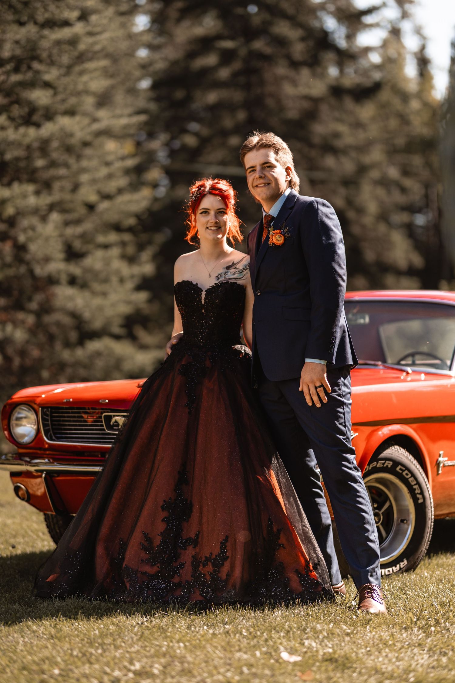 Couple poses by vintage red Ford Mustang, woman in black gothic wedding gown, man in navy suit, forest backdrop.