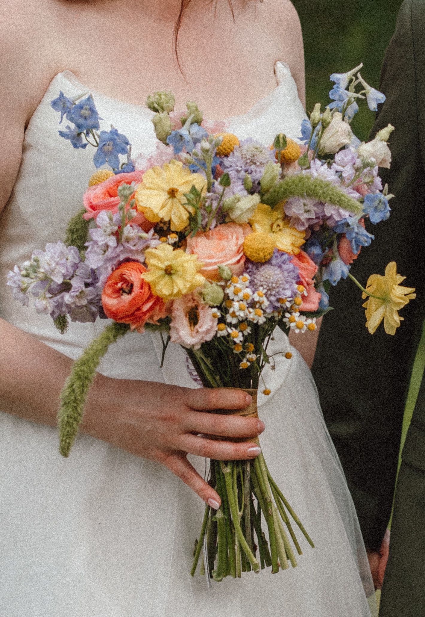 Bride holding a colorful wildflower wedding bouquet with coral, yellow, blue, and lavender blooms in a white gown.